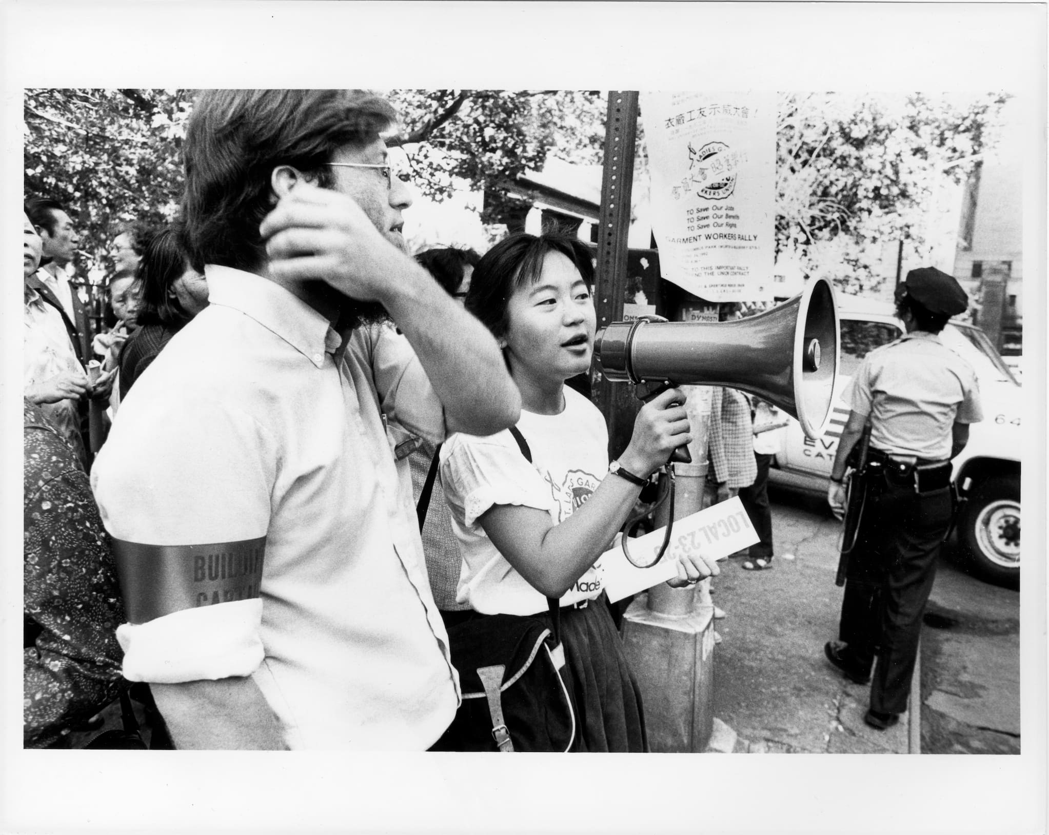 Young woman speaks into megaphone at outdoor rally. Signs and people are in background.