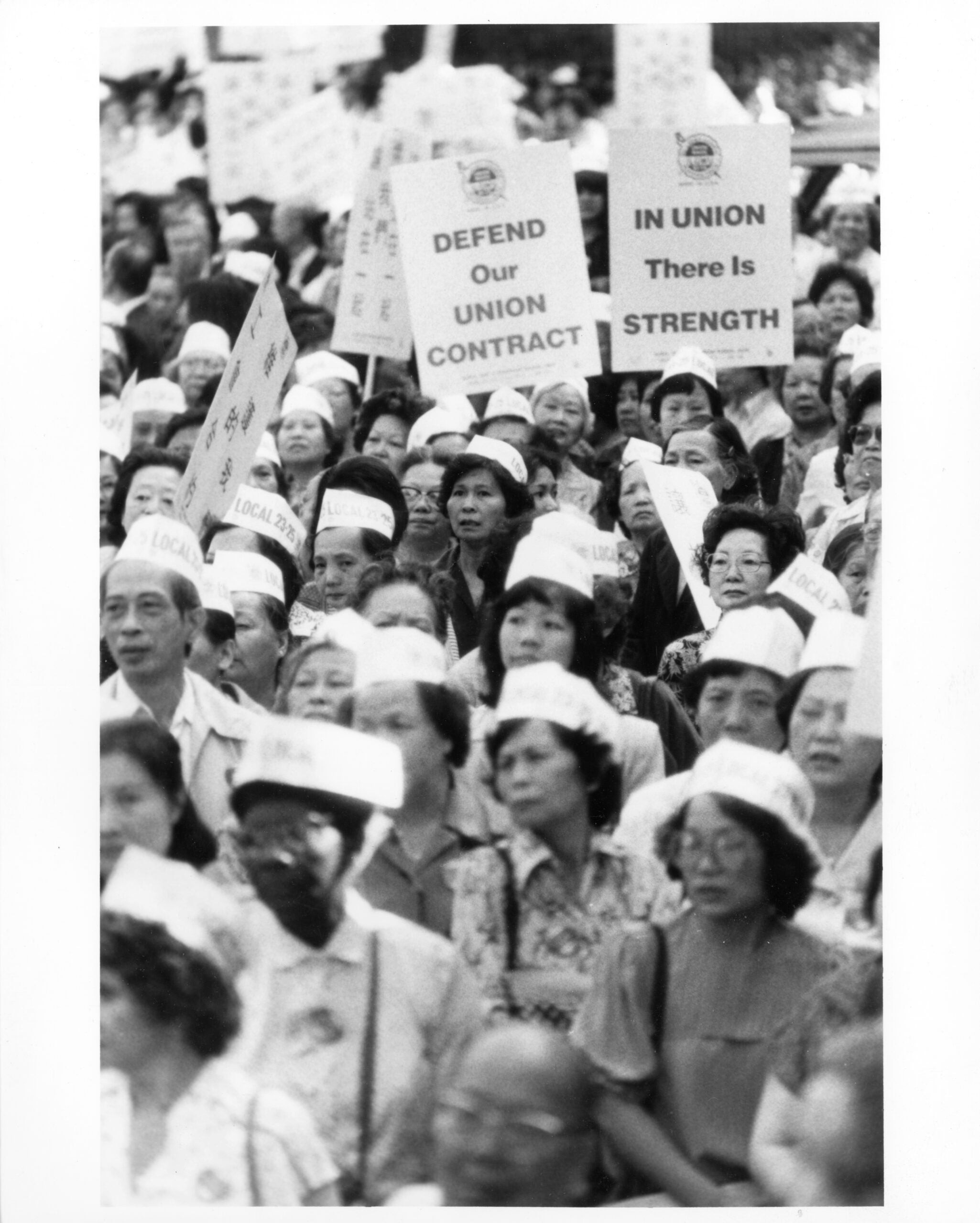 Crowd of people gather at outdoor rally. Signs reading "In Union There Is Strength" and "Defend Our Union Contract" are raised above onlookers.