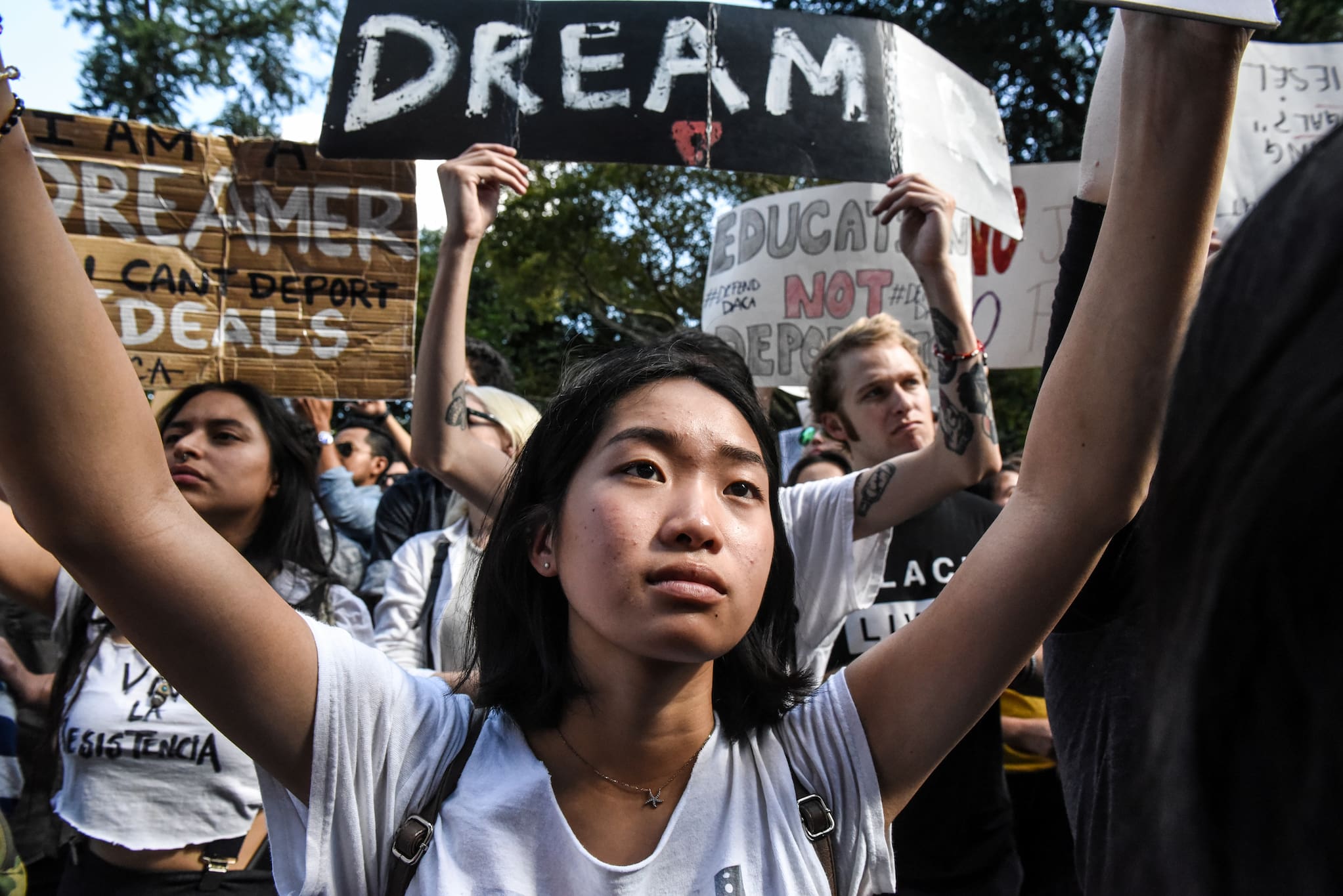 A young Asian American woman holds up a sign above her head at an outdoor rally. Behind her, other protestors hold signs that read "Dream."