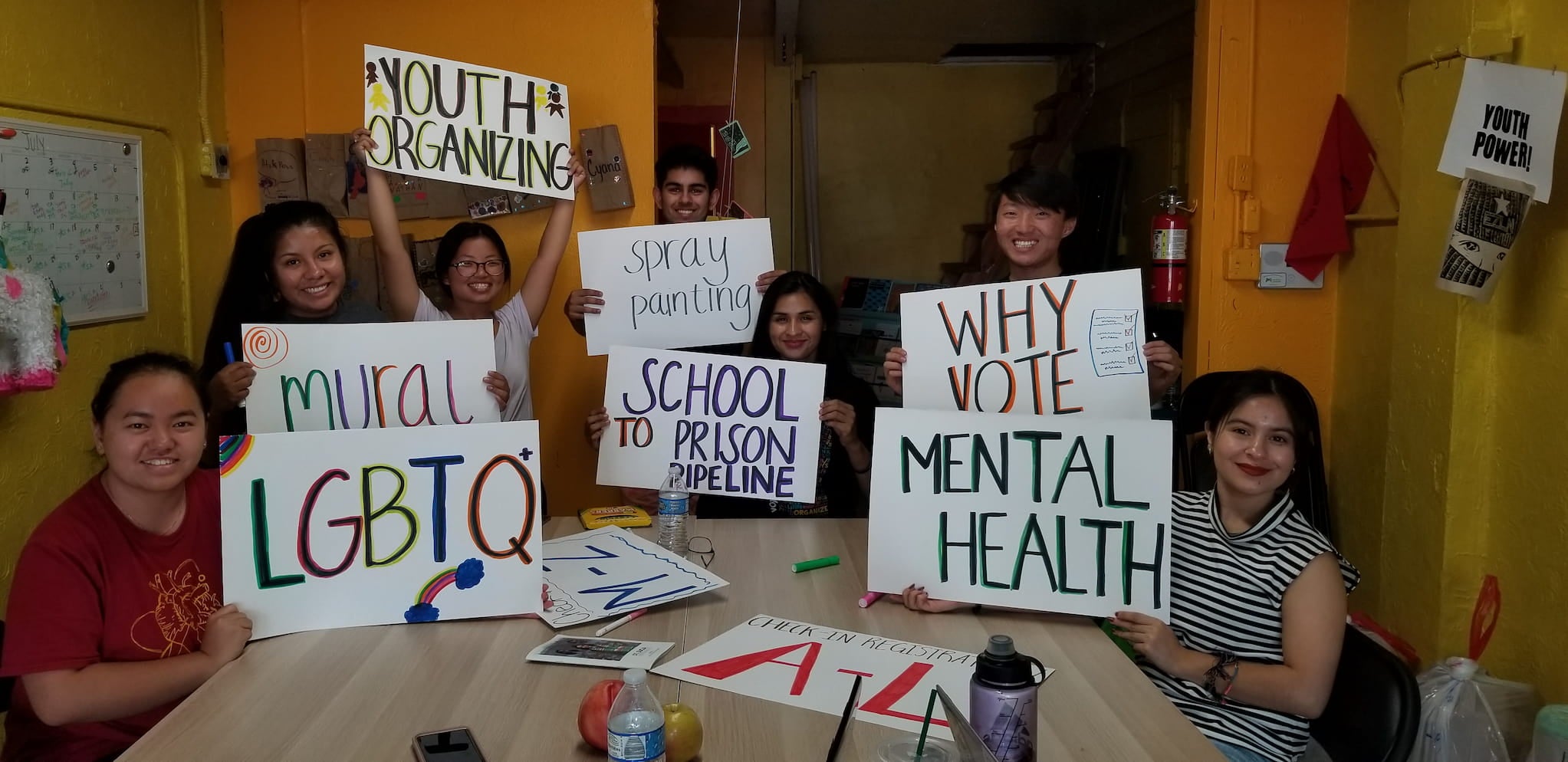 Seven young adults sit around a large, indoor table. They hold up hand made signs about various issues that affect their communities.