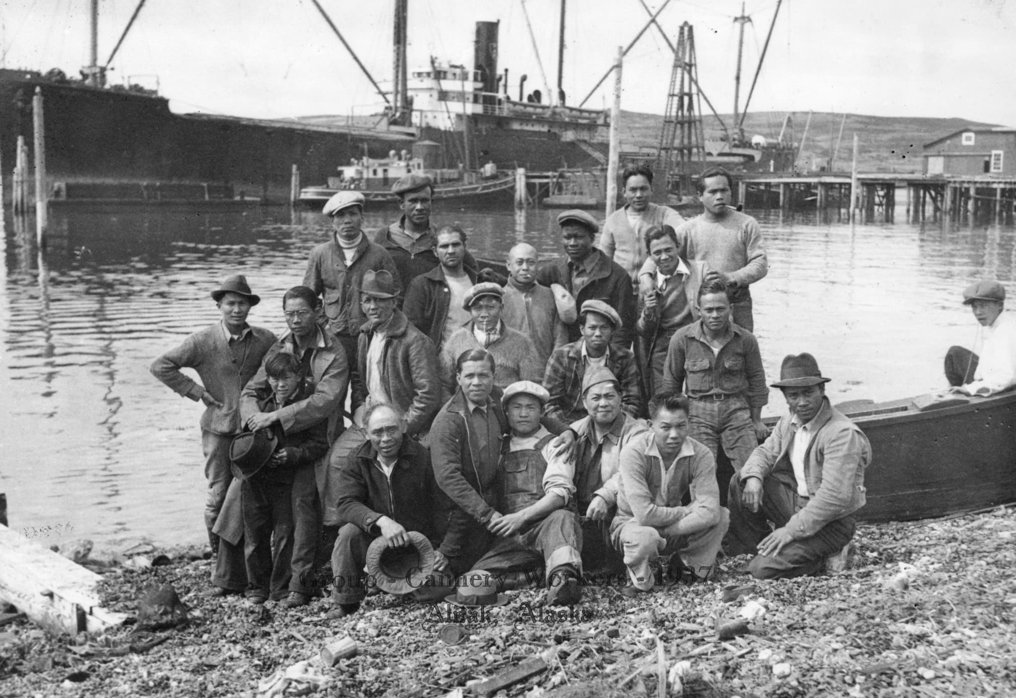 A group of Filipino cannery workers pose on the shoreline in front of the docks.