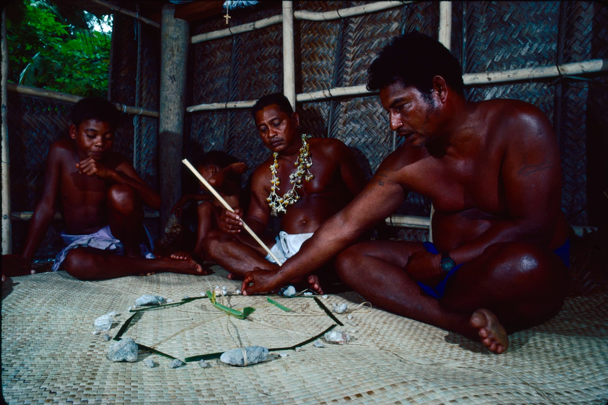 Mau Piailug sits cross-legged and points to a diagram on woven mat. He teaches navigation to two kids and a man seated next to him.