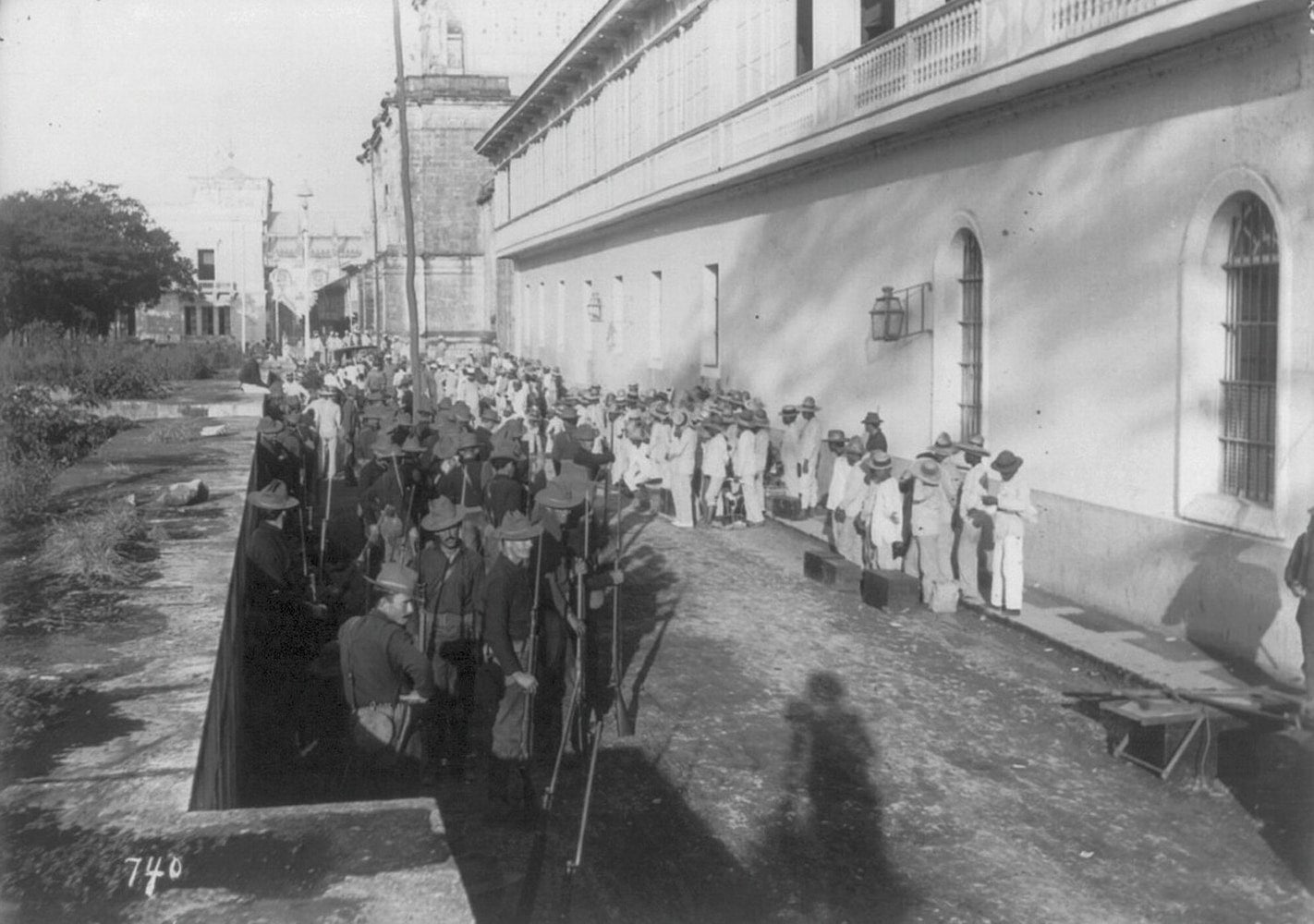 Two columns of people are shown on the outskirts of a town. One column is soldiers holding rifles. The other column is people weaing hats and white clothing.