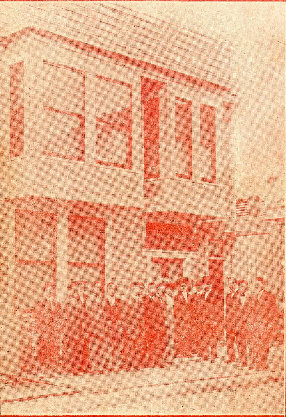 Red color cast photo: A group of men in suits pose outside of a two-story building.