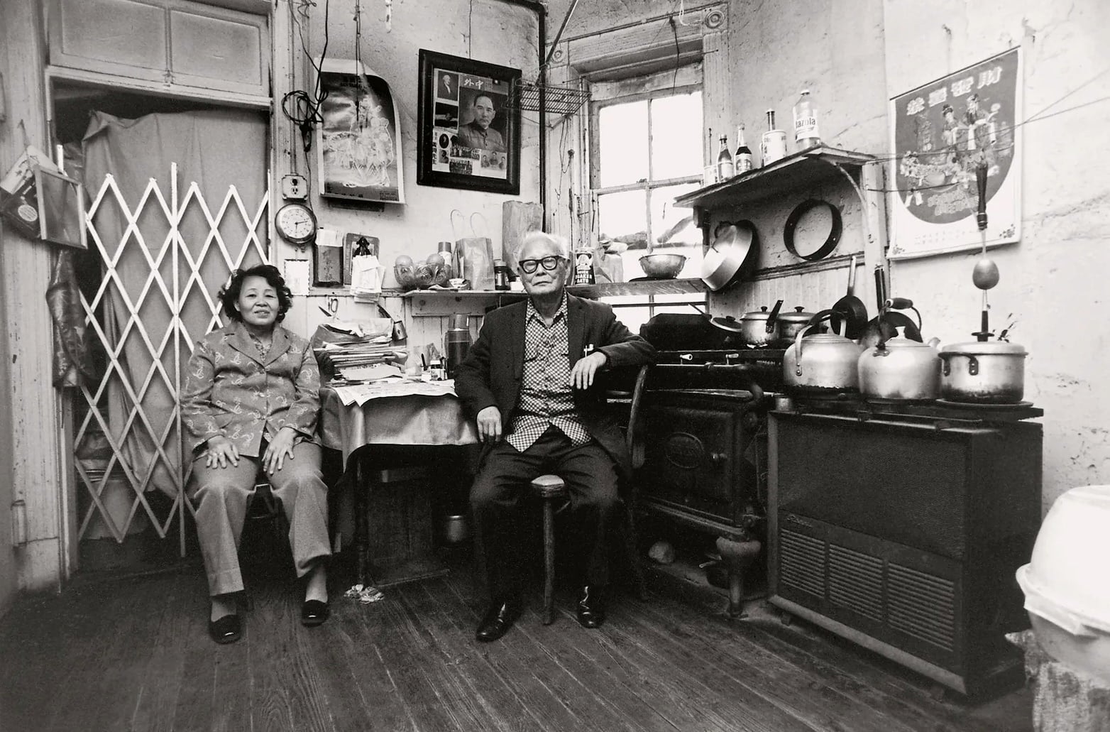 An elderly Asian couple in a kitchen. Pots and pans, stacks of papers, and grocery bags cover every surface. Asian art hangs on the wall.