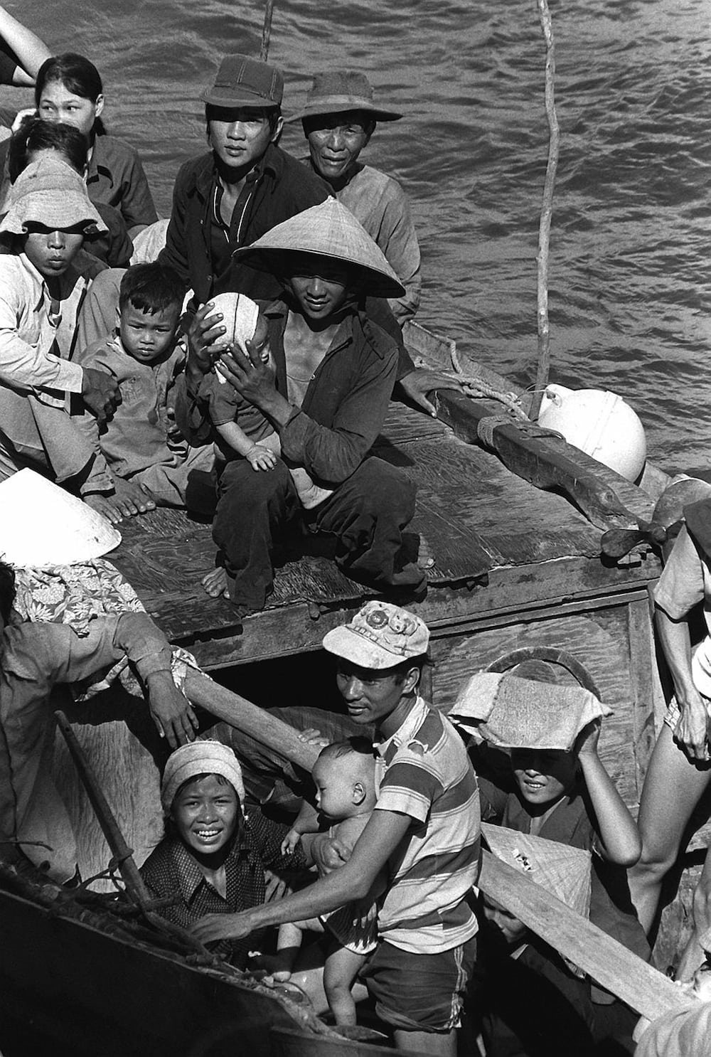 Refugees crowd into a boat. A man in the center sits on the roof of the boat holding a baby.