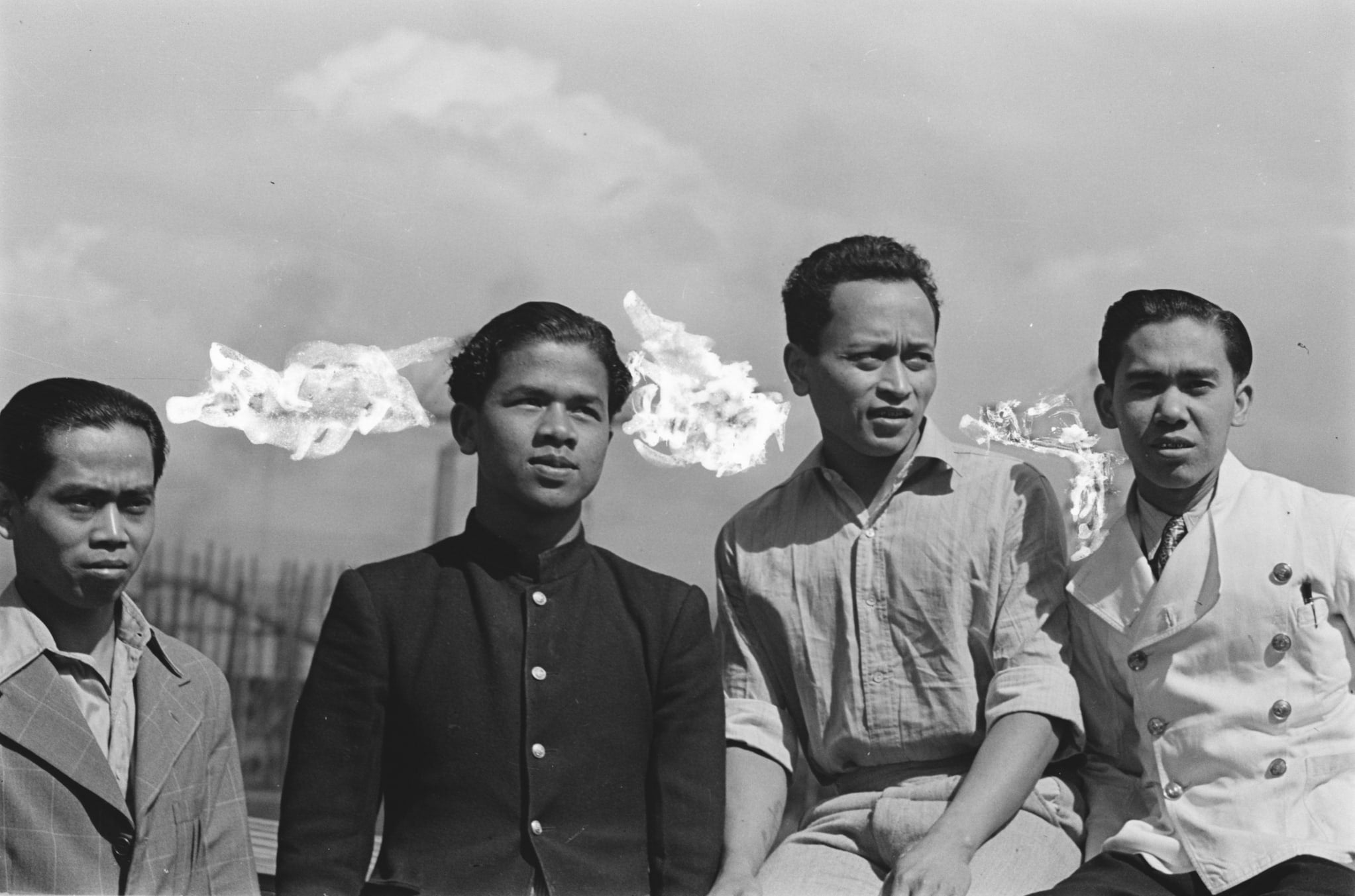 Four young men dressed in jackets and button-down shirts pose in front of a factory.