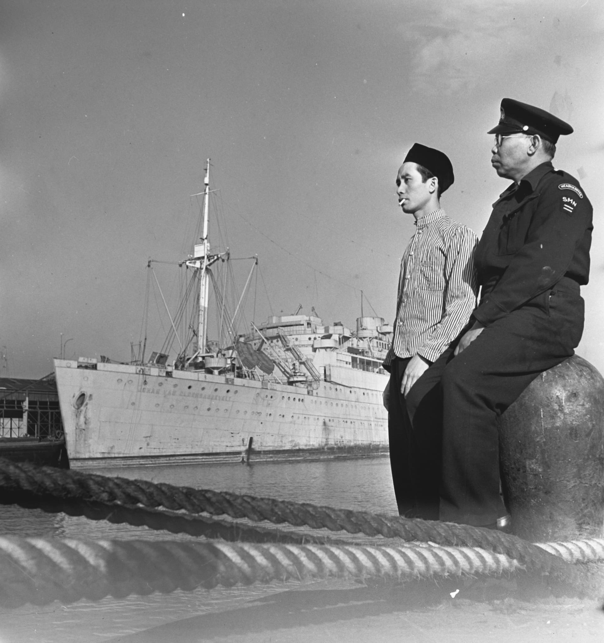 A couple of dock workers lean against a post on the dock. A military ship floats in the background.
