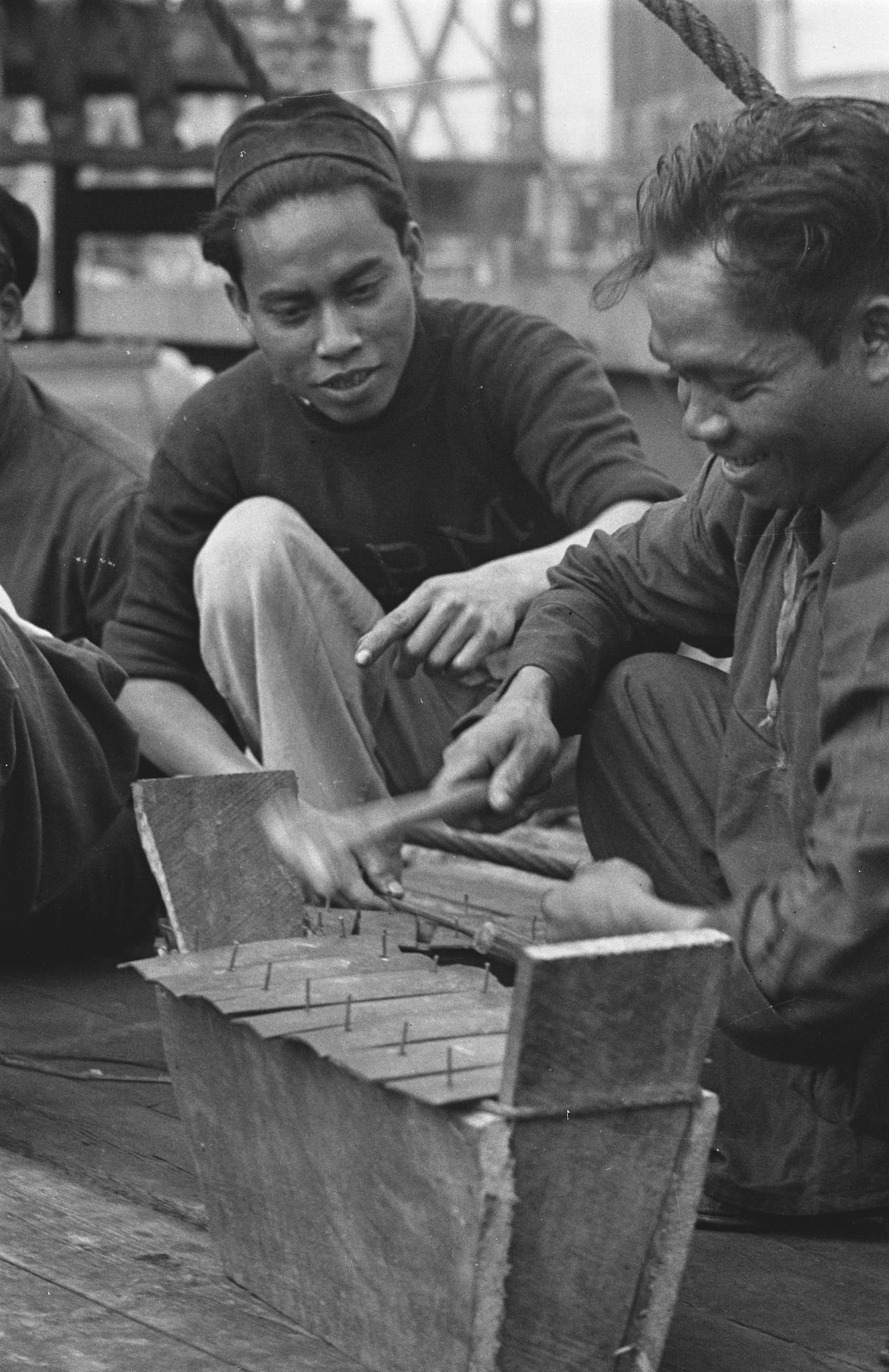 A couple of men smile as they use a hammer to create an instrument that looks like a xylophone on a raised box.