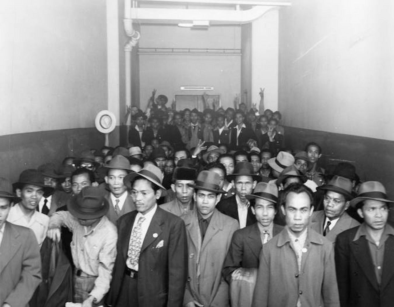 A crowd of men fill a long hallway. Some men have their arms in the air.
