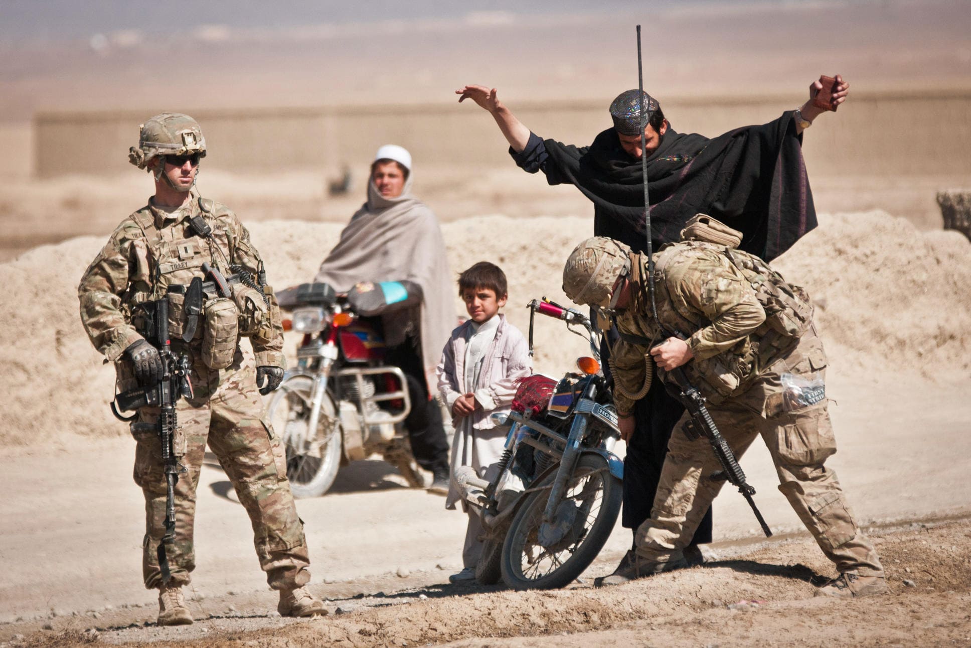 A U.S. soldier searches an Afghan man with raised arms. Another U.S. soldier and a boy stand next to them. Another man on a motorbike awaits his turn.