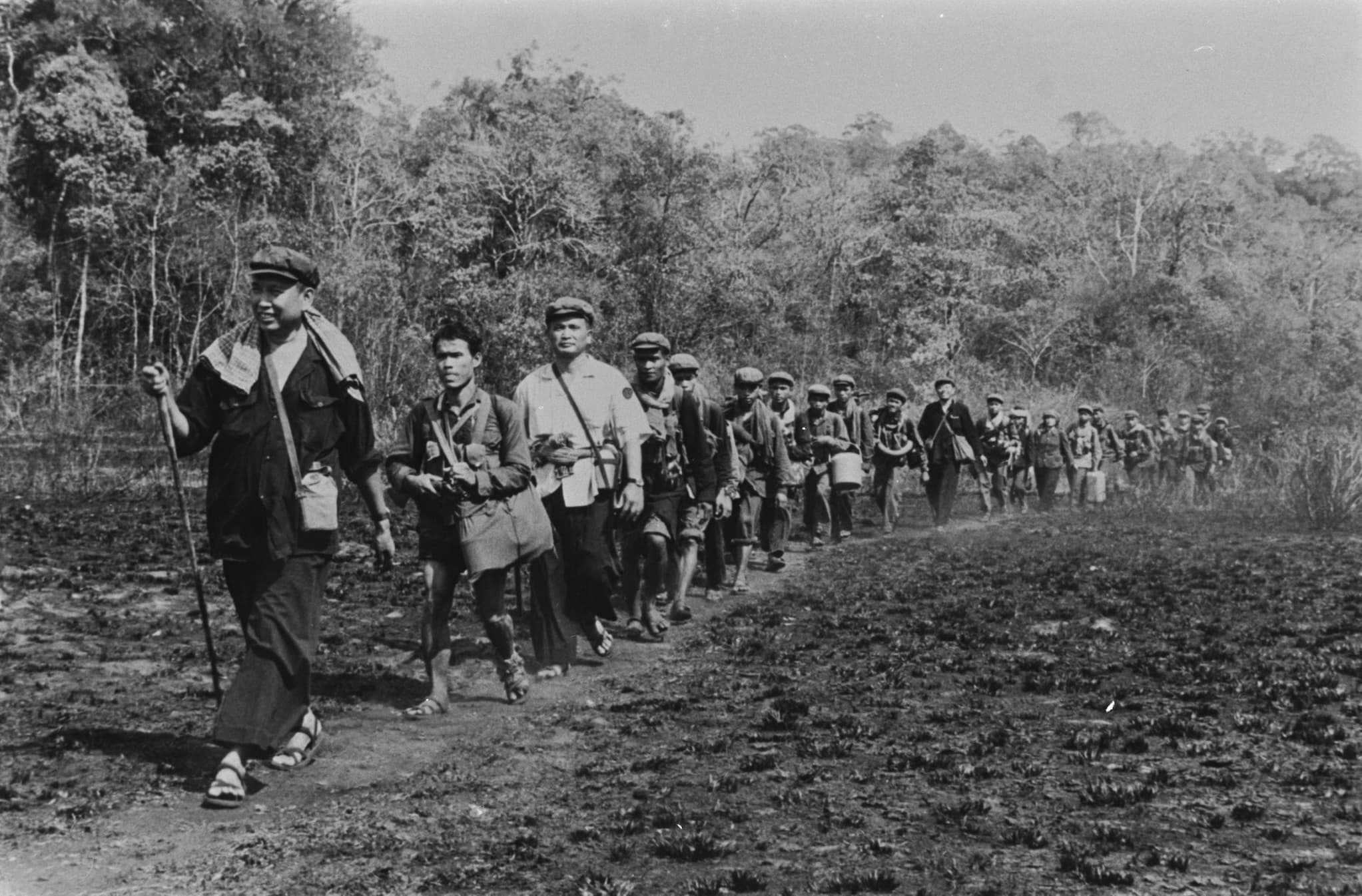 Pol Pot in a black shirt and pants, sandals, and a flat cap carries a stick as he leads as members of the Khmer Rouge along outdoor path.