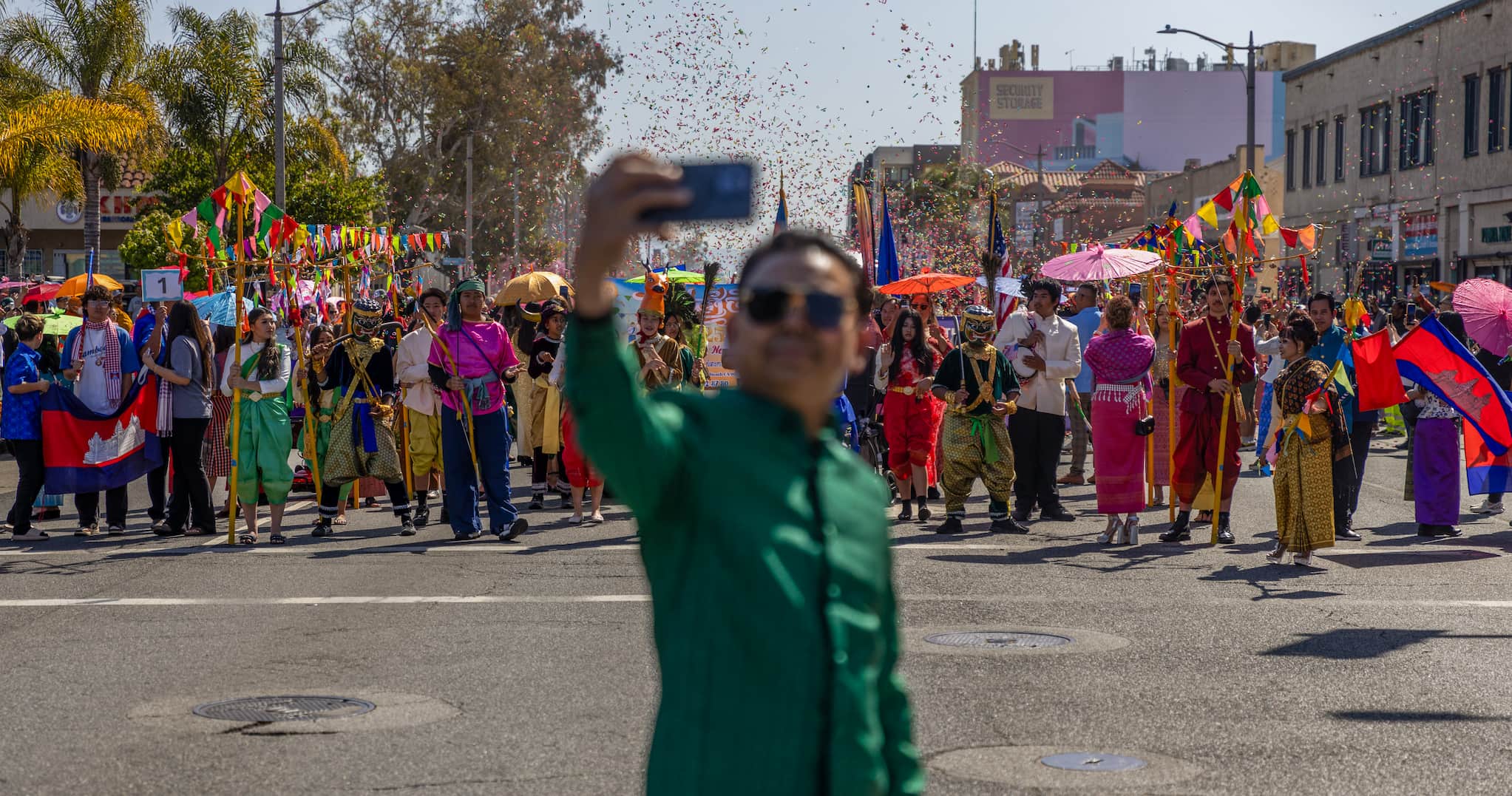 A man takes a selfie in front of the parade in Cambodia Town. Confetti fills the air above the paraders dressed in brightly colored attire.