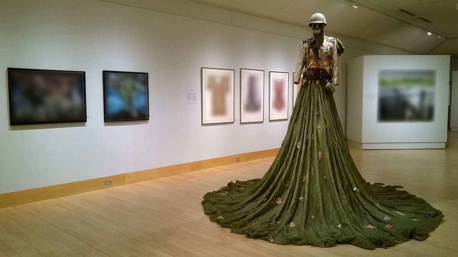 Art piece of a statue with a skull head wearing a soldier's helmet and a dress made out of a military parachute displayed in a gallery.