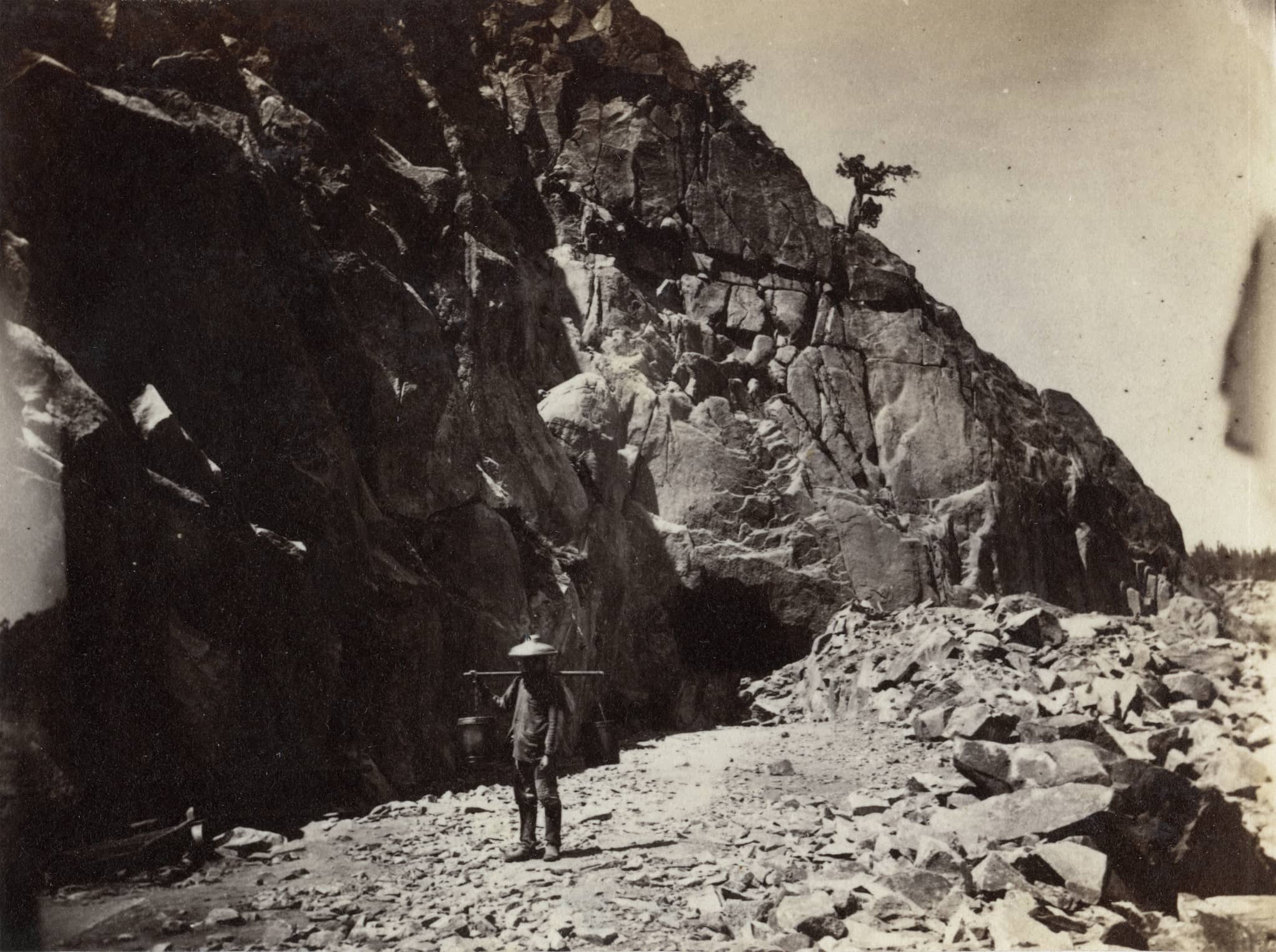 A Chinese man carrying a pole with a bucket on either end over his shoulders standing between a mountainside and piles of rubble.