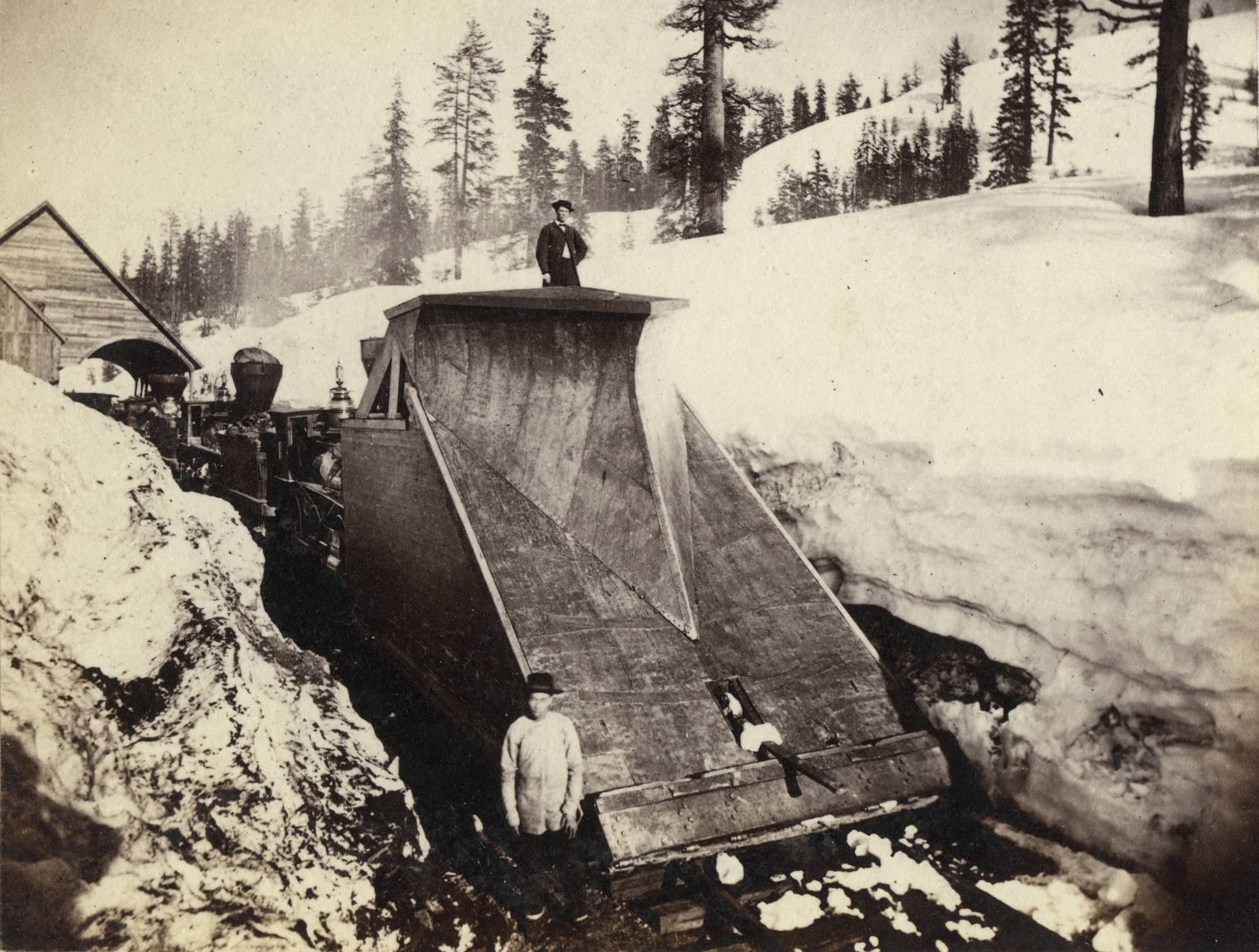 Chinese man stands atop a snow plow affixed to the front of a train while a Chinese worker stands at the bottom as it paves a path through deep snow.