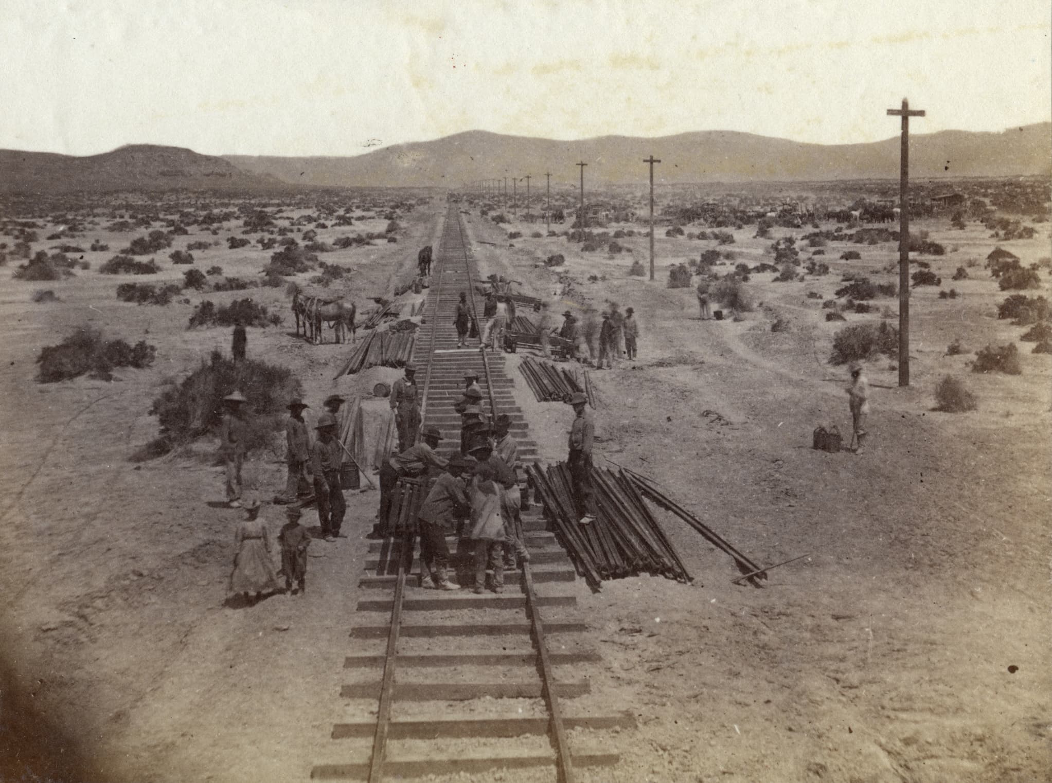 A group of Chinese railroad workers laying tracks and ties in the desert leading into the mountains. Two women walk alongside the tracks.