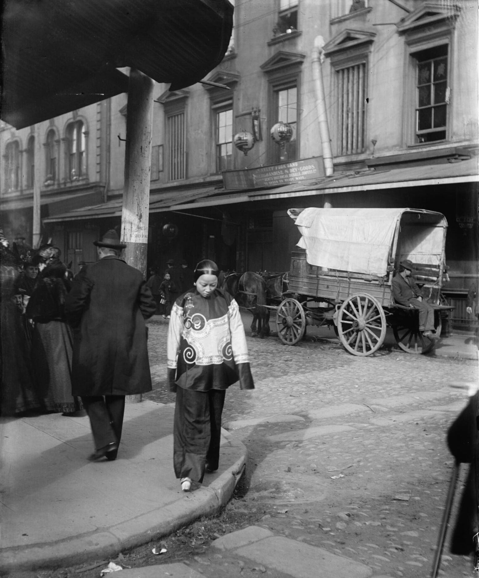 Chinese woman in holiday shanku with chanzu bound feet walking away from a group of white people in Chinatown. Horse-drawn wagon parked on the street.