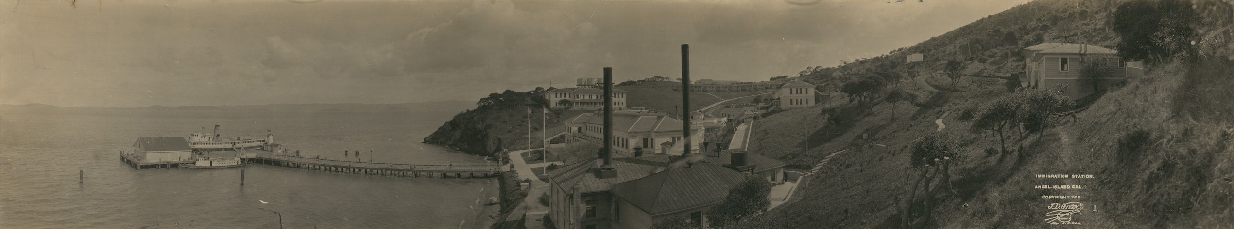 Boathouse and a dock with a long walkway that leads to several buildings posted on a hillside on the edge of the island with two tall smokestacks.