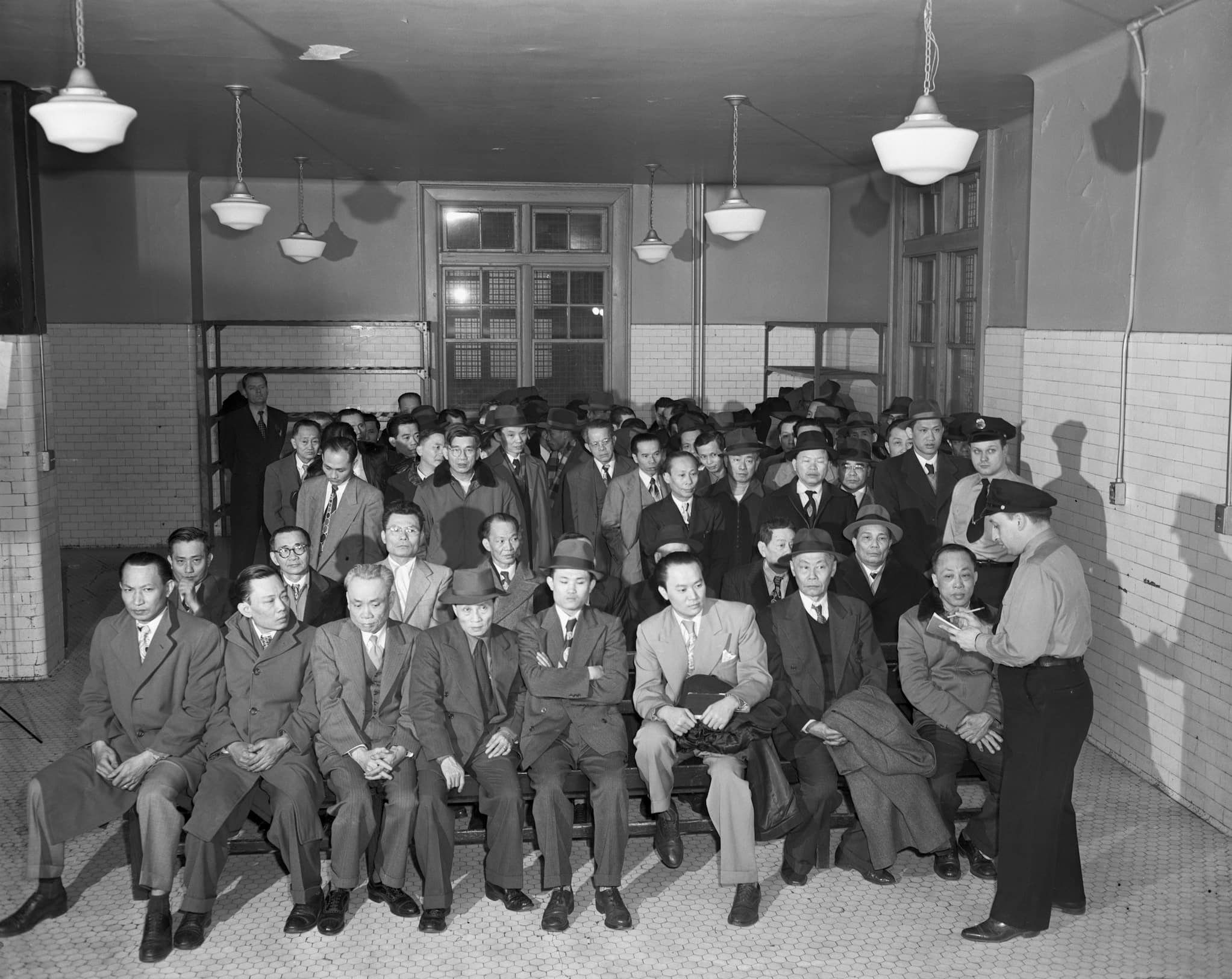 On Ellis Island, two white immigration officers interrogate a group of Chinese immigrant men suspected of being Communists or deserting seamen.
