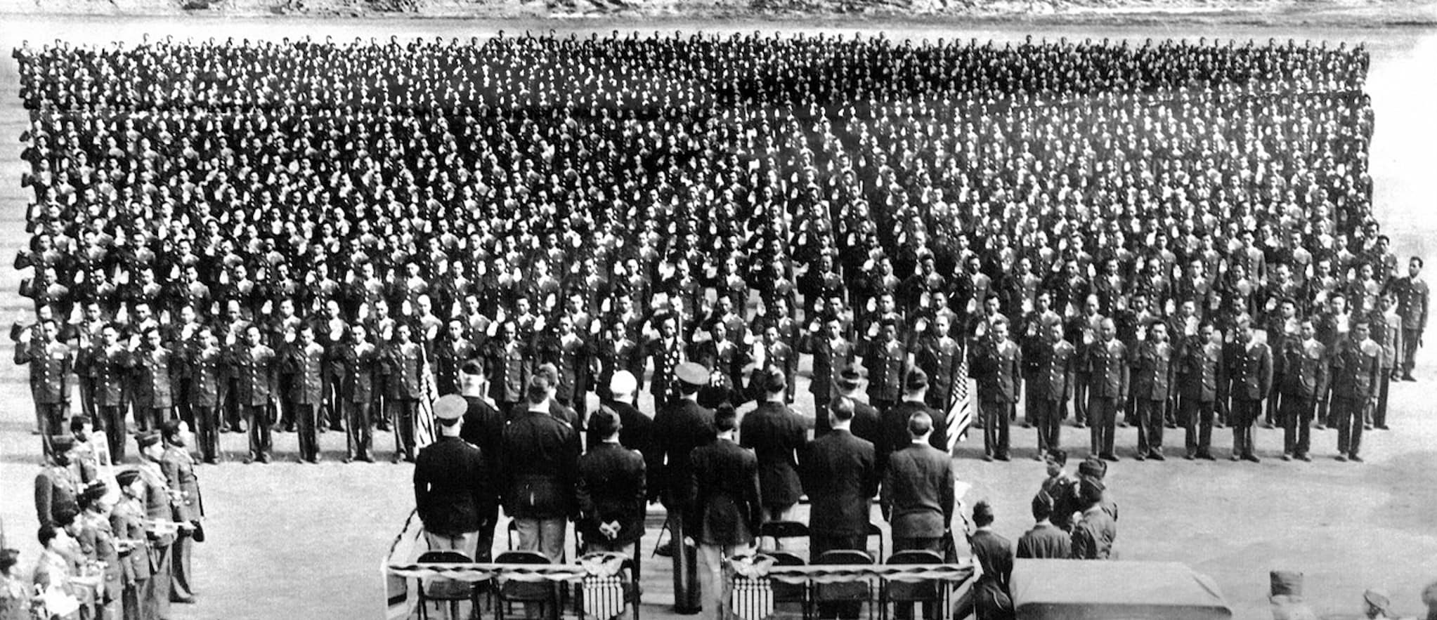 One thousand uniformed soldiers stand in multiple rows during outdoor ceremony. Group of men stand at the head of the crowd, flanked by U.S. flags.