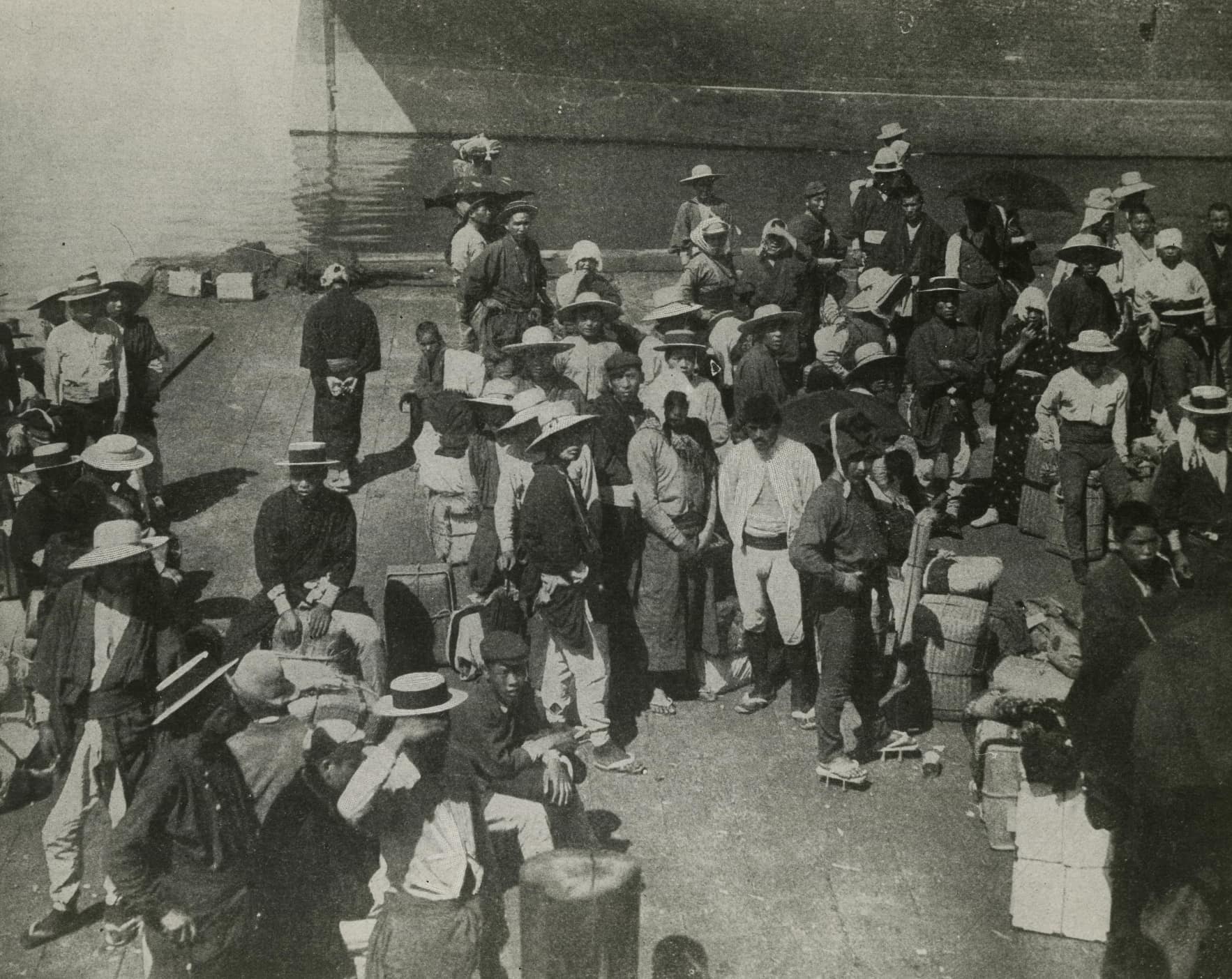 A crowd of Japanese immigrants dressed with hats, casual wear, and their luggage stand at the docks. Bottom of large ship appears in the background.