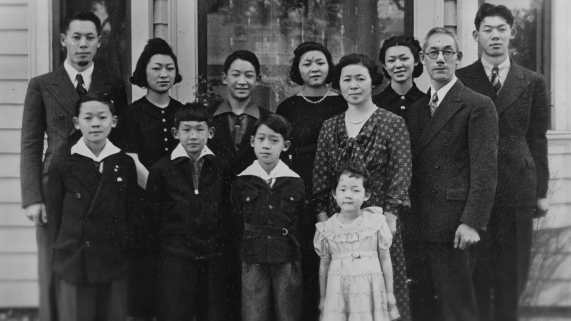 A portrait of the Japanese American Uno family. Four children and eight adults dressed in dark colors pose outside for picture.