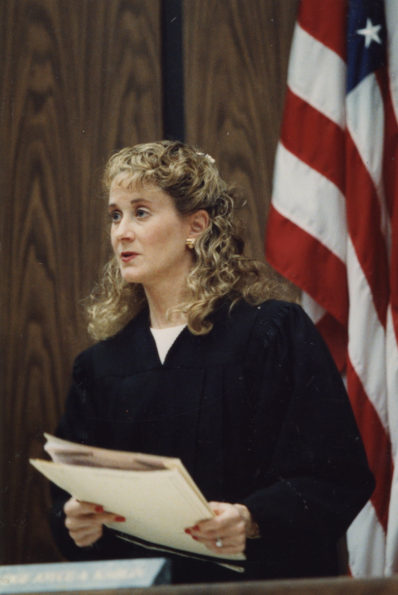 Joyce Karlin, a white woman in judge's robe, stands in courtroom in front of a U.S. flag. She holds a case file in her hands.