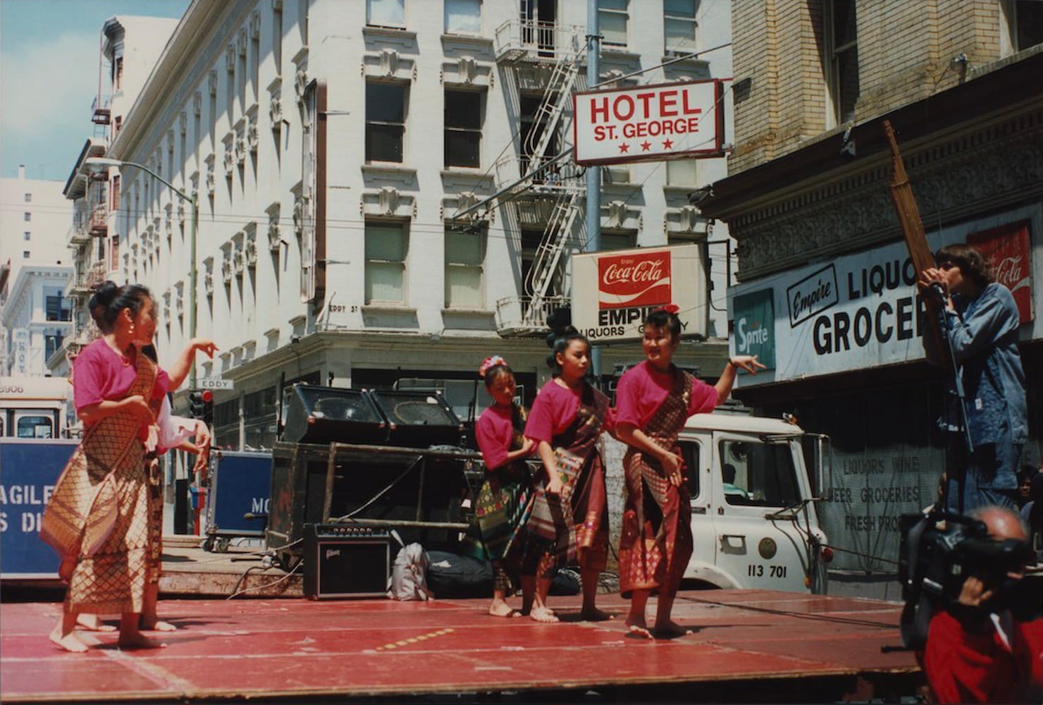 Young girls, all dressed in matching shirts, skirts, and shawls, perform on an outdoor stage. On the right a man plays a Lao mouth organ on stage.