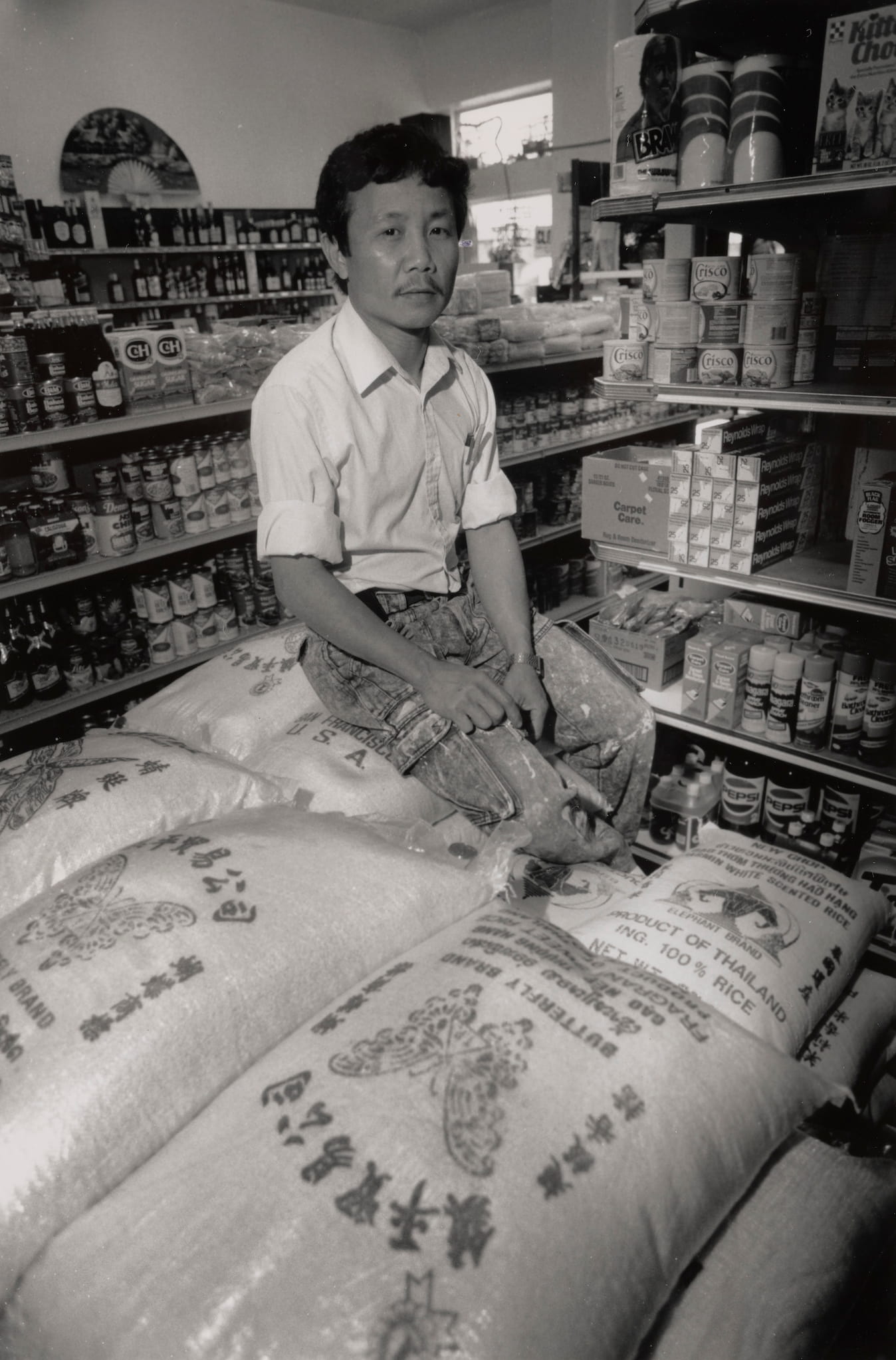 Khamchanh Louanphixay, a man dressed in a button down shirt with the sleeves rolled up, sits on a stack of bagged rice in the middle of his store.