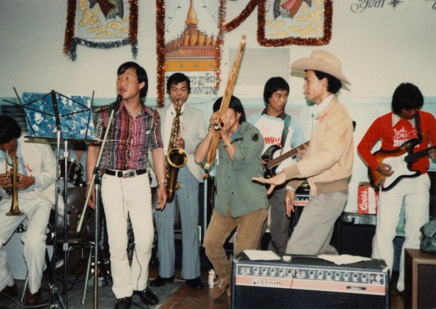 Eight Laotian men celebrate Lao New Year by singing, dancing and playing musical instruments in front of festive Lao decorations.