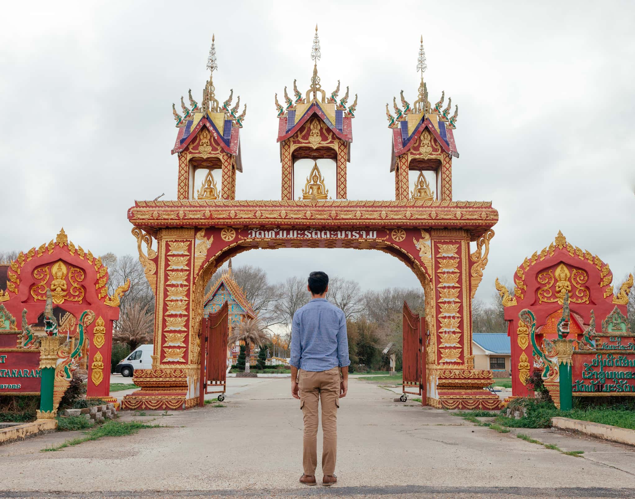 Lao immigrant Phanat Xanamane, dressed in a buttoned down shirt and pants, stands facing the entrance of Wat Thammarattanaram with its ornate gate.