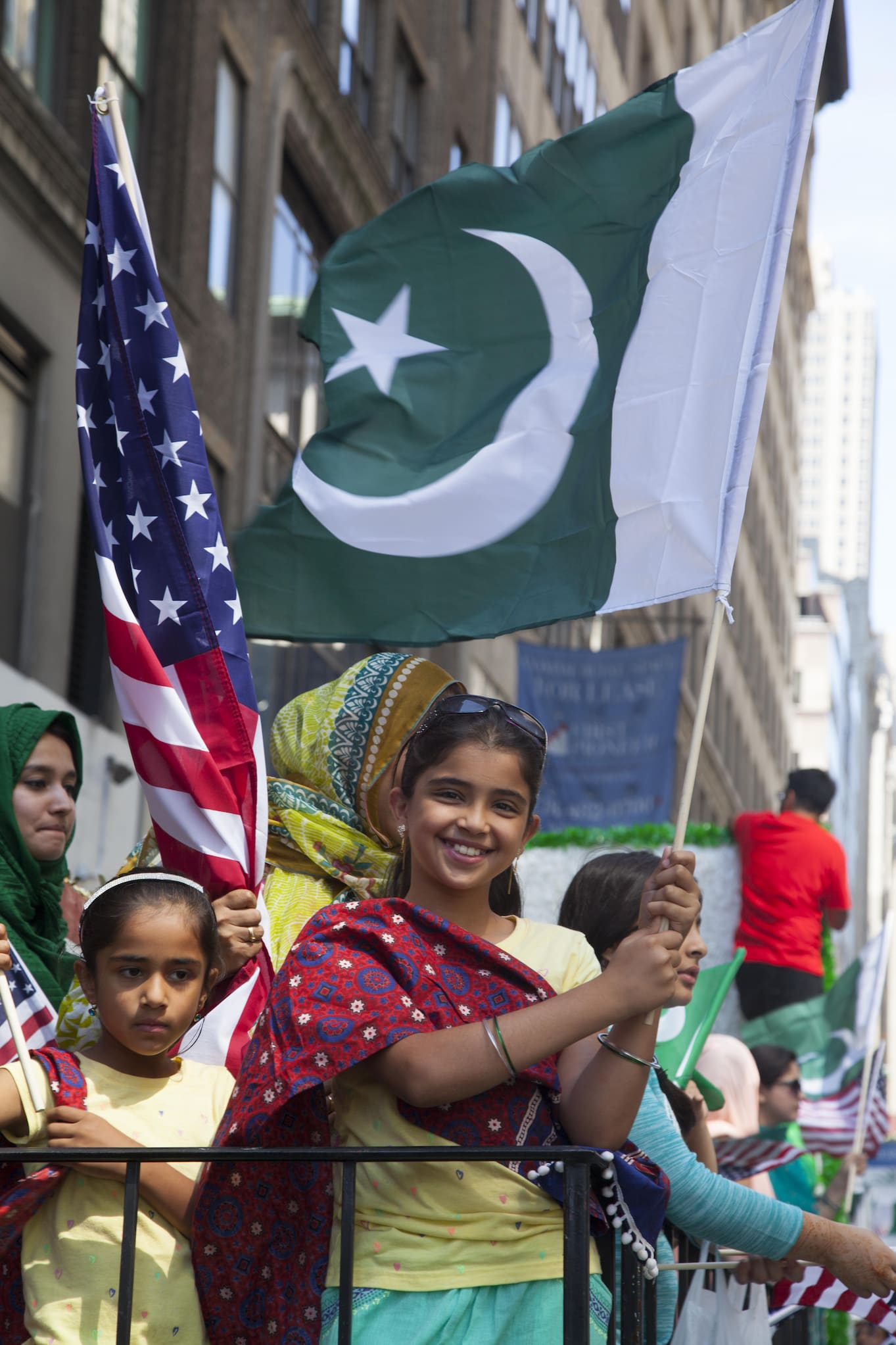 Kids hold American and Pakistani flags on the side of a parade.