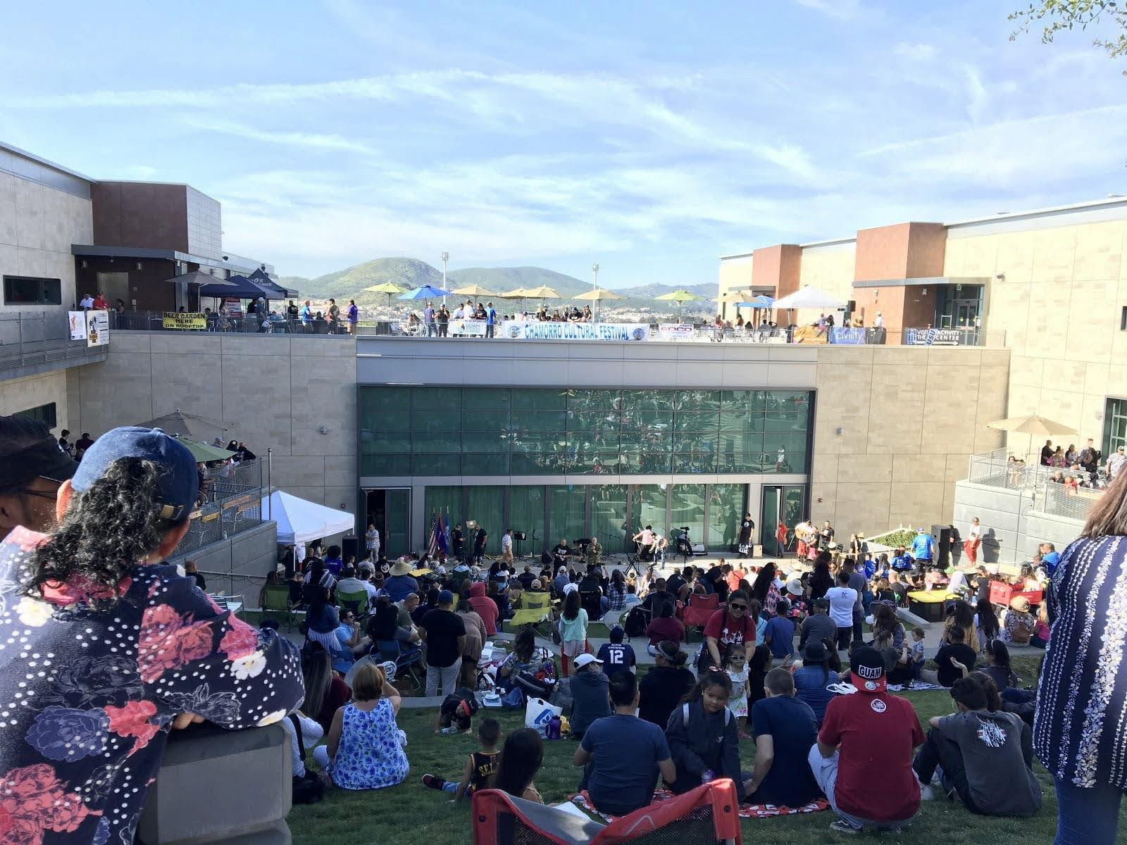At outdoor festival, a crowd gathers on the grass in front of a building at CSU San Marcos. They watch people set up on a makeshift stage.