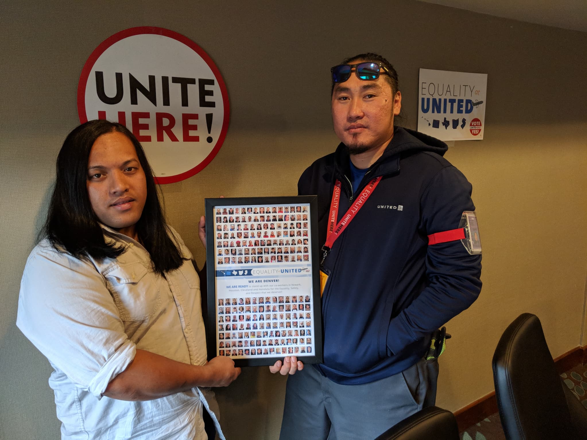 Two United employees hold a framed poster that reads "Equality United" between them. The pair stands in front of a sign that reads, "Unite Here!"