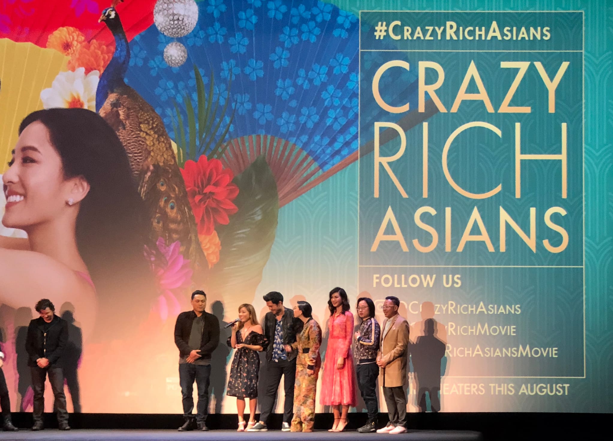 The cast of Crazy Rich Asians stand in a row on stage. Behind them, a large screen displays the film's key art.