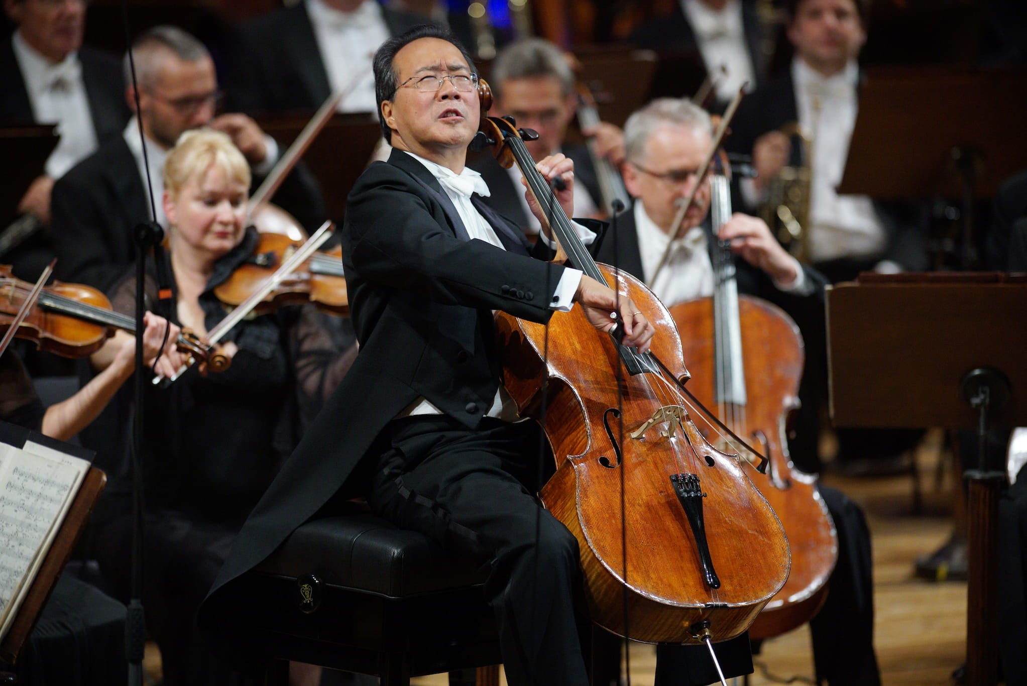Cellist Yo-Yo Ma, dressed black suit jacket, performs on stage. Other orchestra members perform behind him.