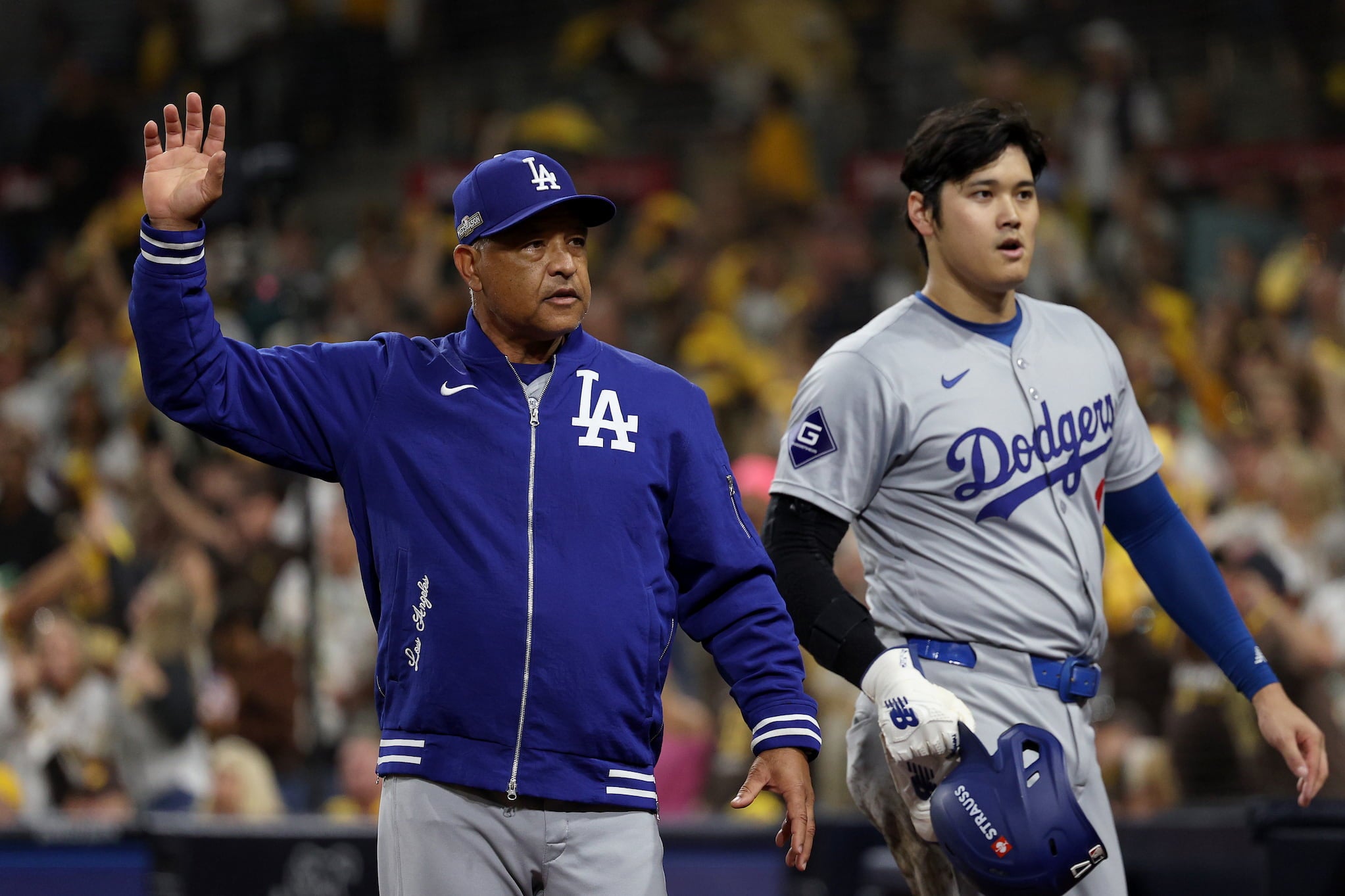 Dave Roberts, dressed in blue Dodgers zip-up jacket, walks with hand raised next to Shohei Ohtani, dressed in gray Dodgers uniform and helmet in hand.
