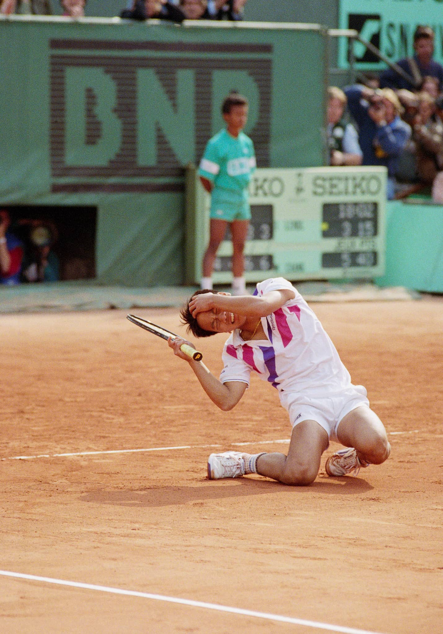 Tennis player Michael Chang, wearing a white uniform and clutching his tennis racket, collapses on the court with a pained expression on his face.