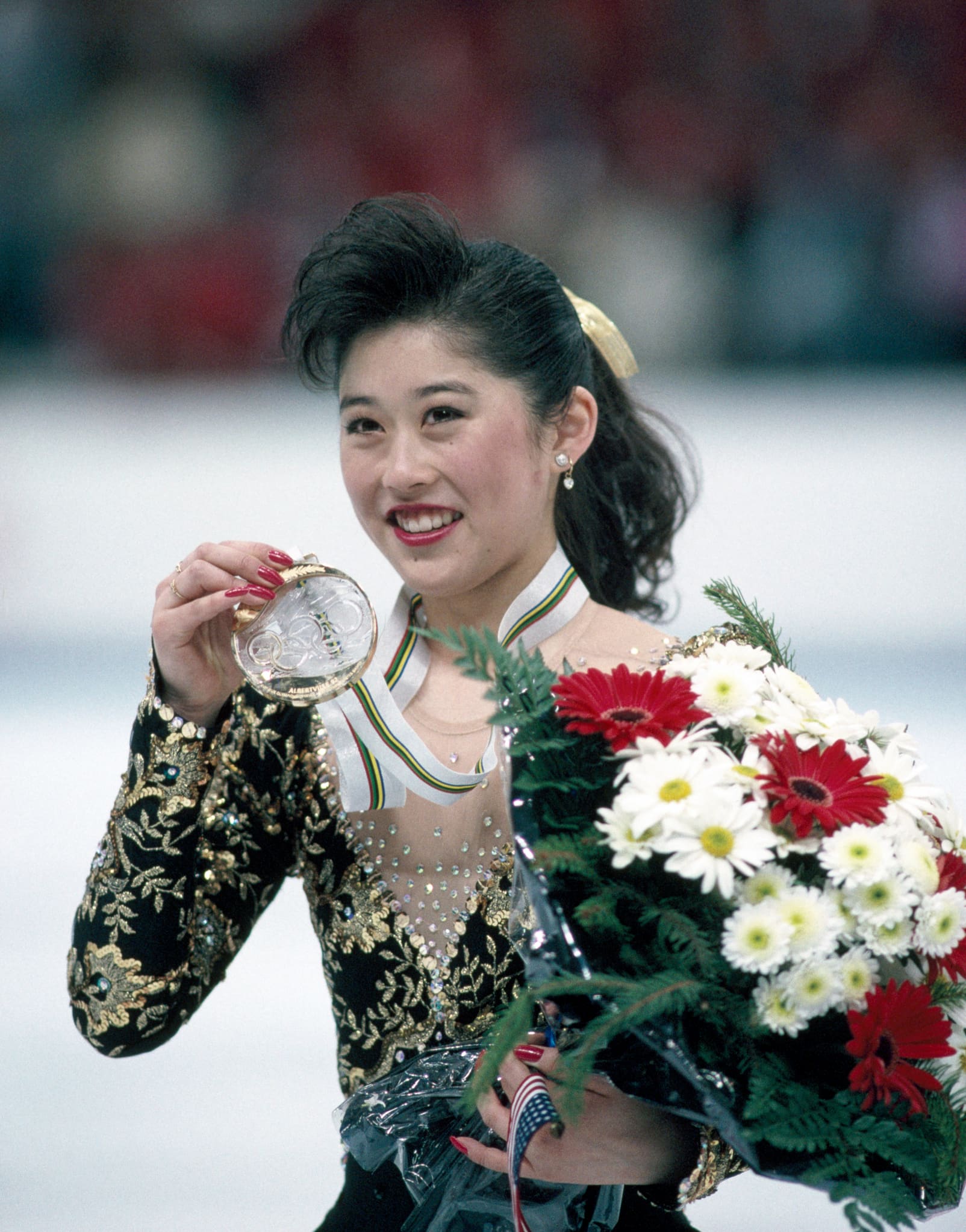Kristi Yamaguchi, dressed in an embellished figure skating dress, holds a large bouquet in one hand and her Olympic gold medal in the other.