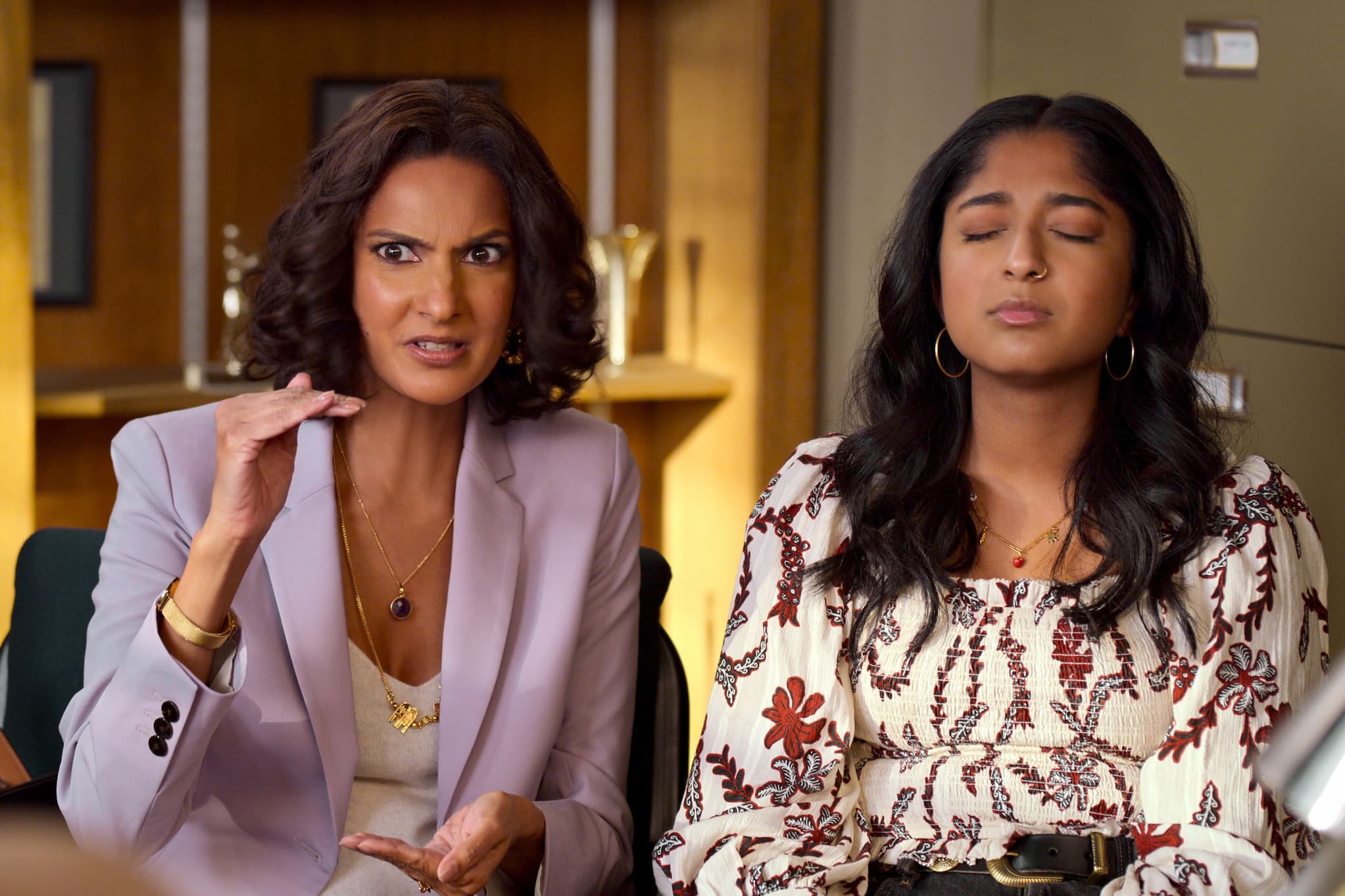 South Asian mother and teenage daughter sit side-by-side in office. The daughter sits with eyes closed while her mother gestures to someone offscreen.