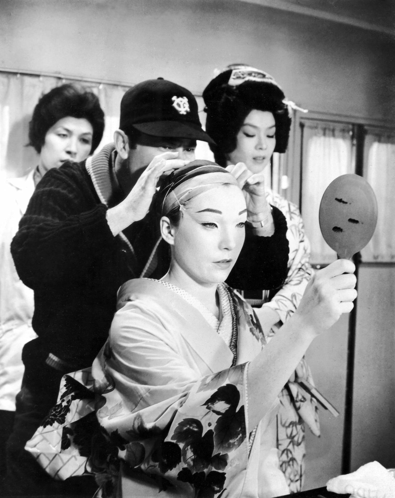 Shirley Maclaine in a make-up trailer wearing a kimono and having her hair styled as two Asian women look on.