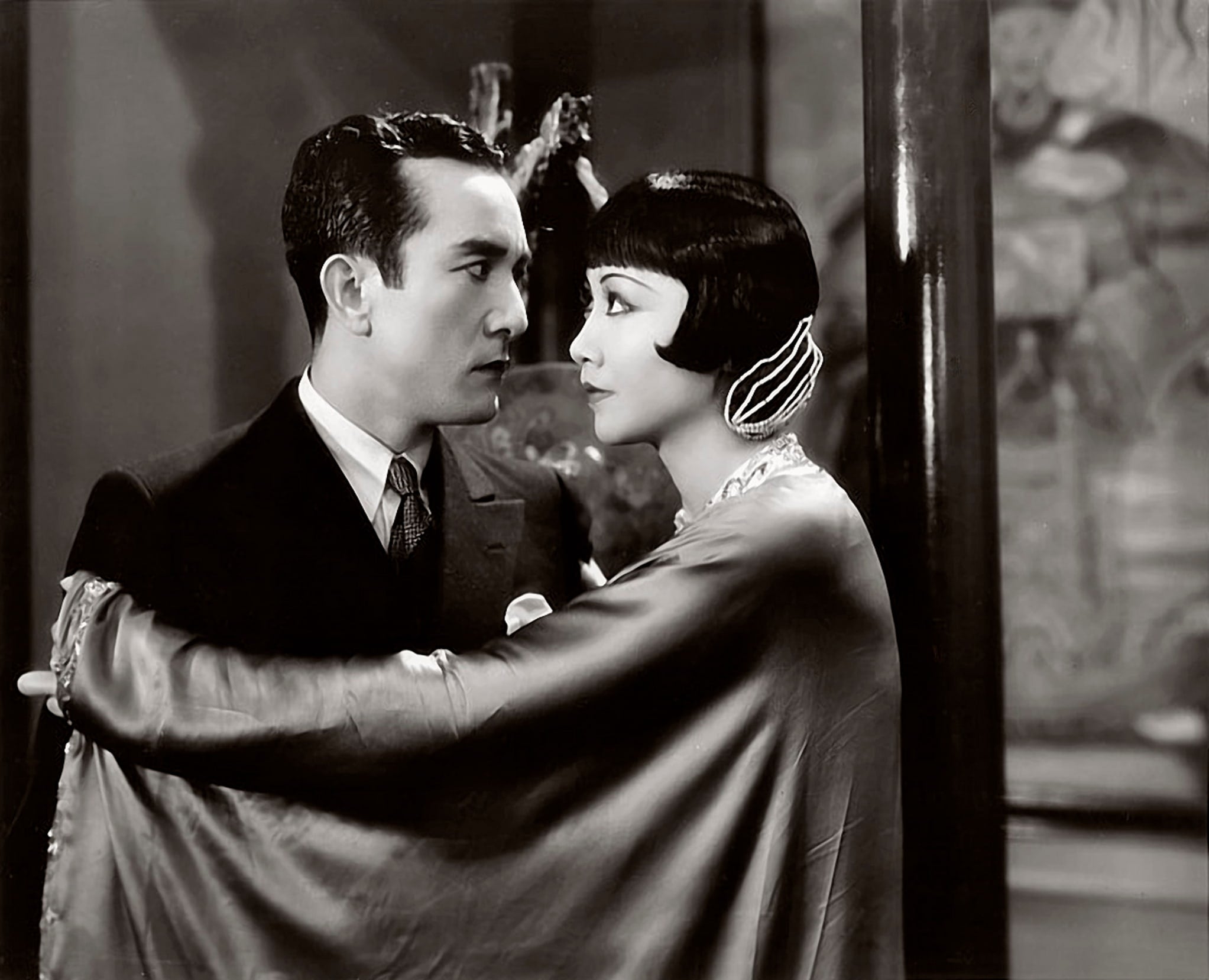 Anna May Wong, Chinese American woman draped in silk clothing, wraps arm around Sessue Hayakawa, Japanese American man in suit and tie.