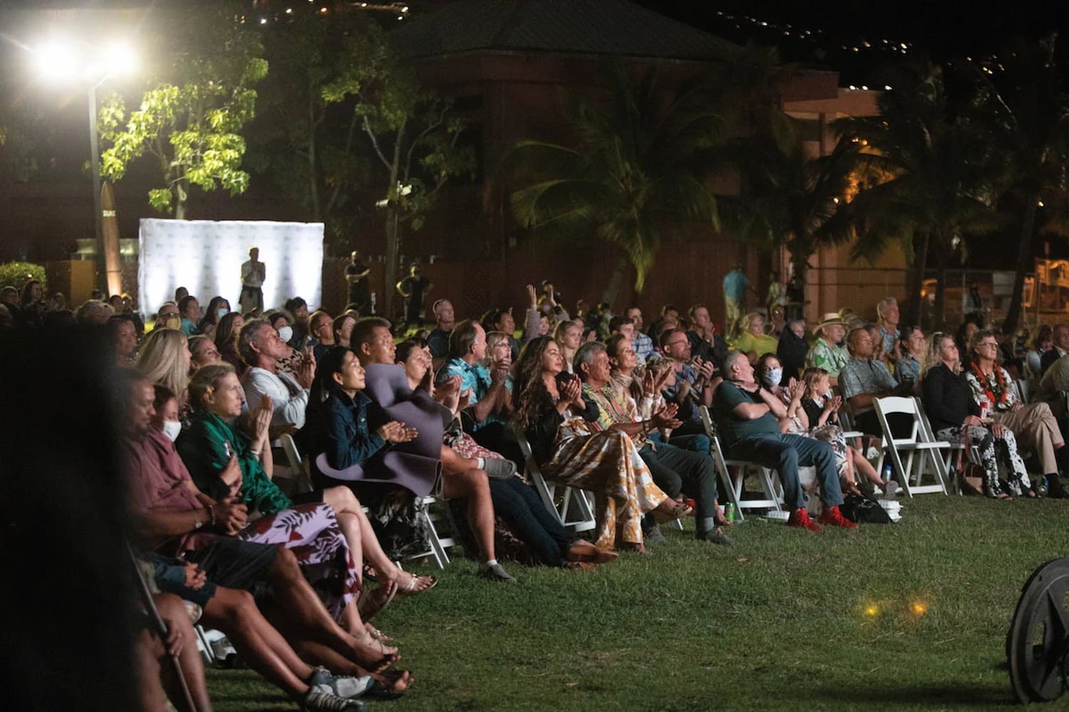 Audience gathers on lawn for night time screening at the Hawaiʻi International Film Festival. People sit in rows on white folding chairs.
