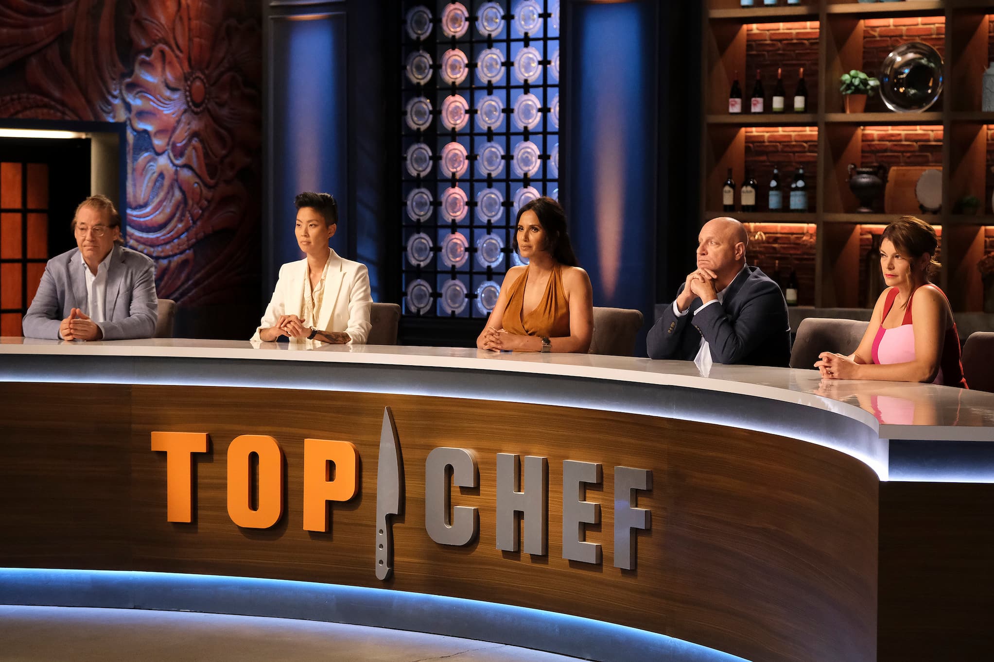 Five people sit at judges' table on Top Chef set. Host Padma Lakshmi, an Indian American woman, sits beside Kristen Kish, a Korean American chef.