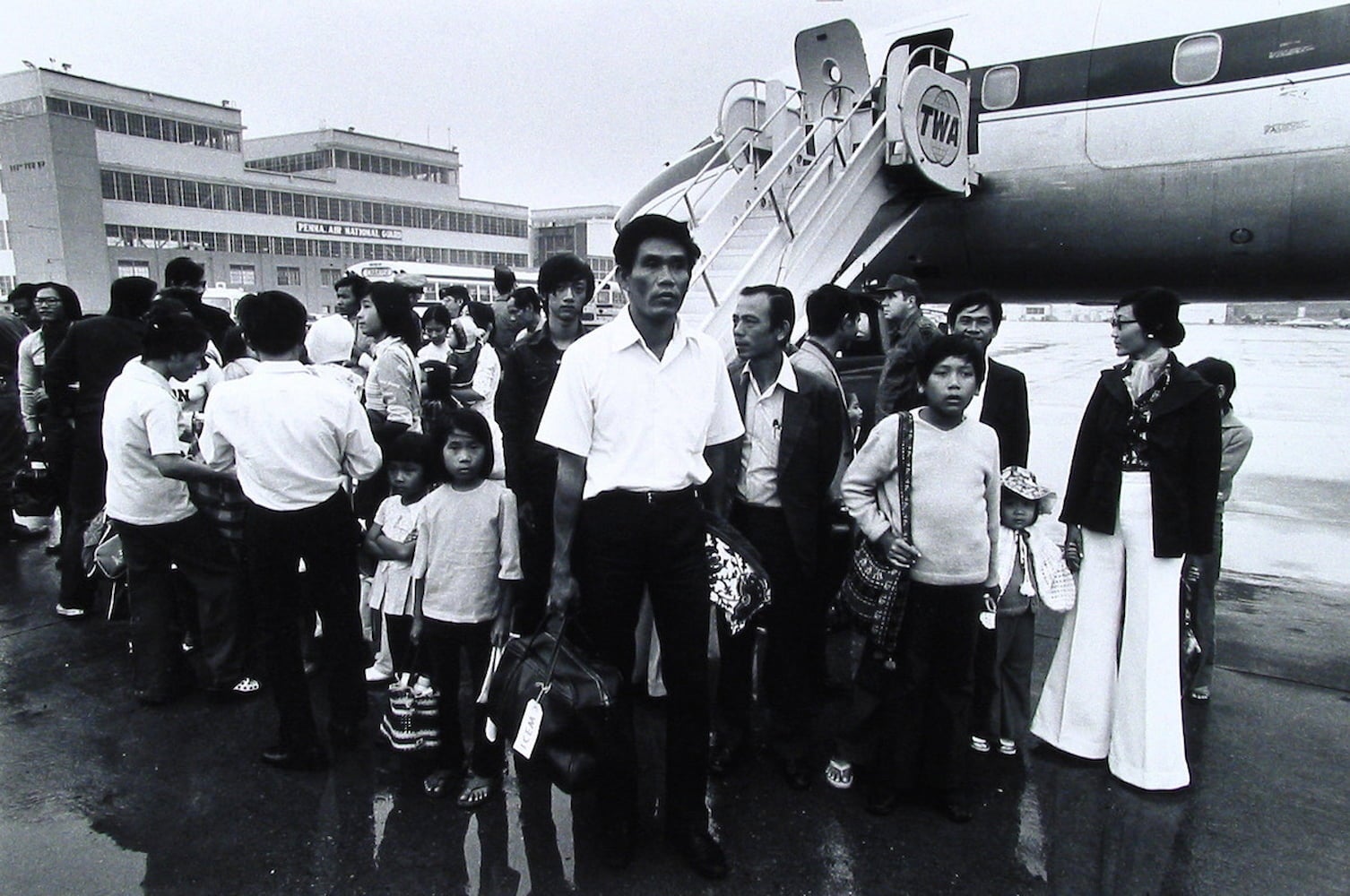 A crowd of Vietnamese refugees, holding their belongings, stand clustered together on the tarmac next to a TWA airplane.
