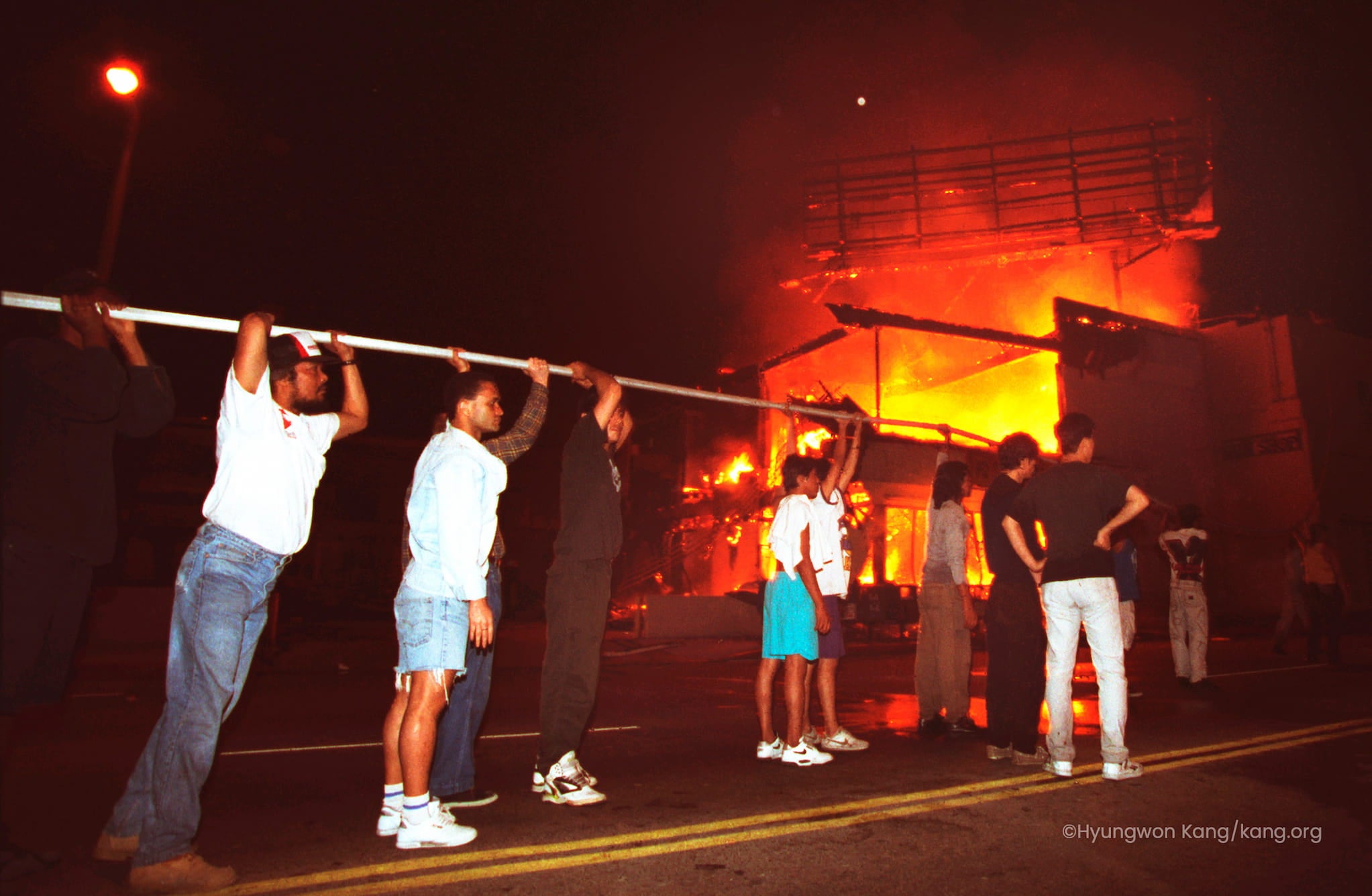In Koreatown at nighttime, fourteen civilians hold up a water hose to extinguish the flames raging through a burning multi-story shopping center.