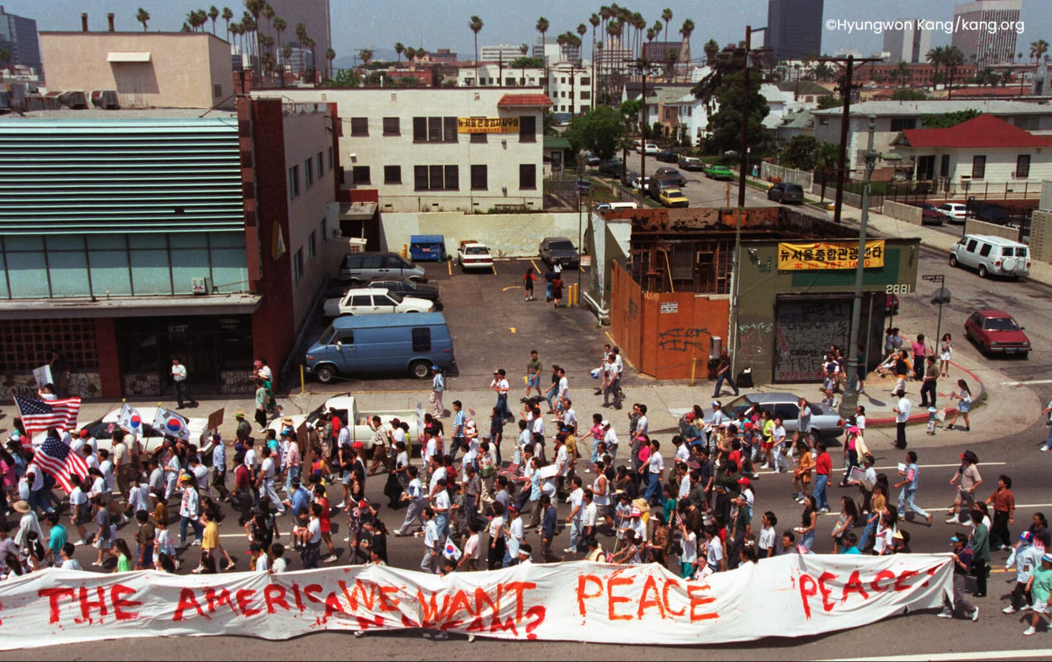 From an elevated view, people march in the street, holding flags and a large banner that spans the crowd. The banner calls for peace.
