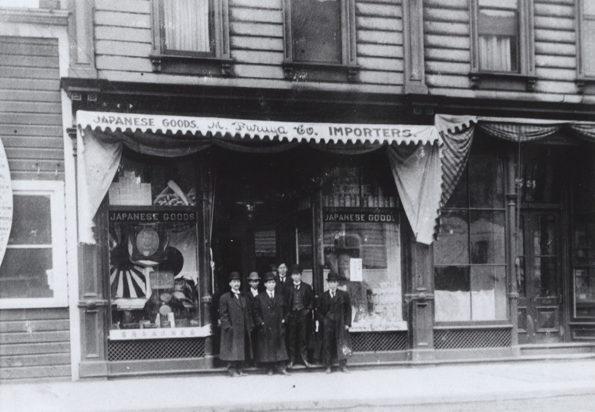 Seven Japanese American men in three piece suits, trench coats, and hats, stand in front of M. Furuya Company. Japanese goods are in window displays.