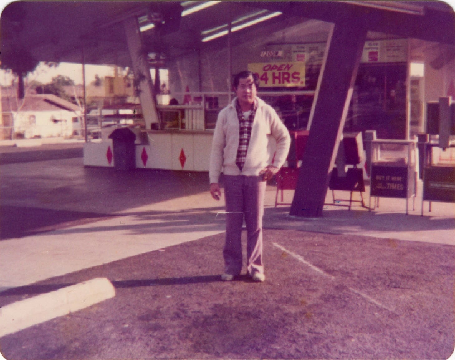 Cambodian American Ted Ngoy, dressed in a jacket, plaid shirt, and long pants, stands in front of a donut shop. Sign behind him reads, "Open 24 Hrs."